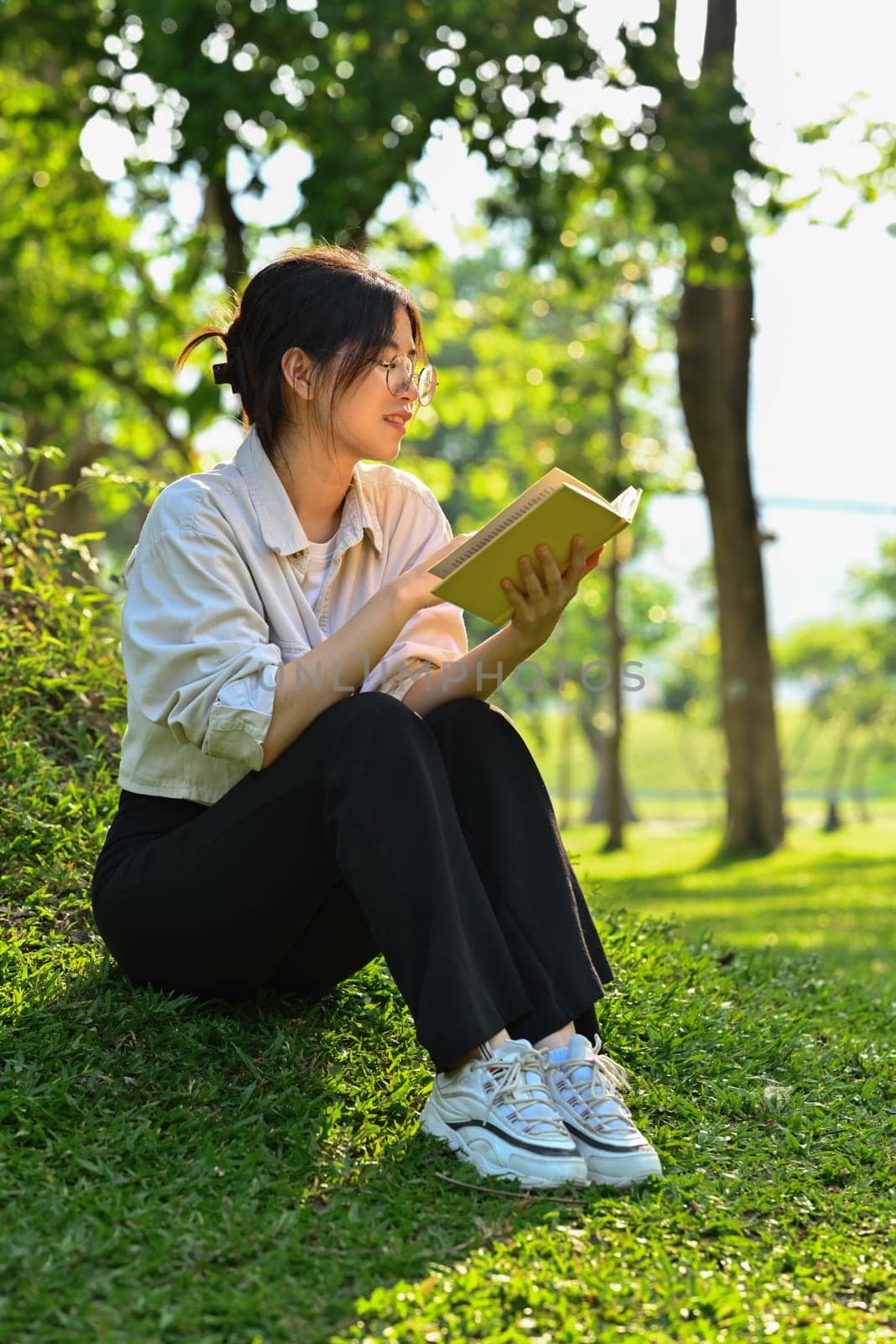 Calm female university student reading book, relaxing on campus lawn. Education, learning and lifestyle concept.