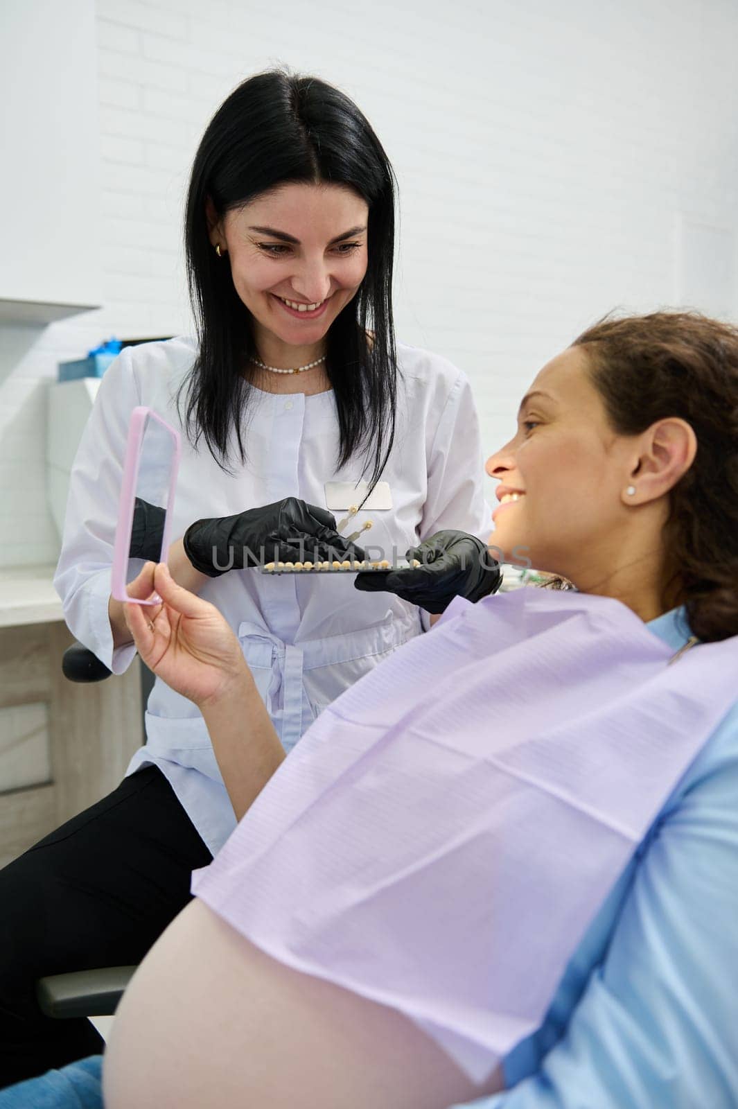 Pregnant woman patient at dental appointment, sitting in dentist's chair, smiling in mirror, discussing with the doctor by artgf