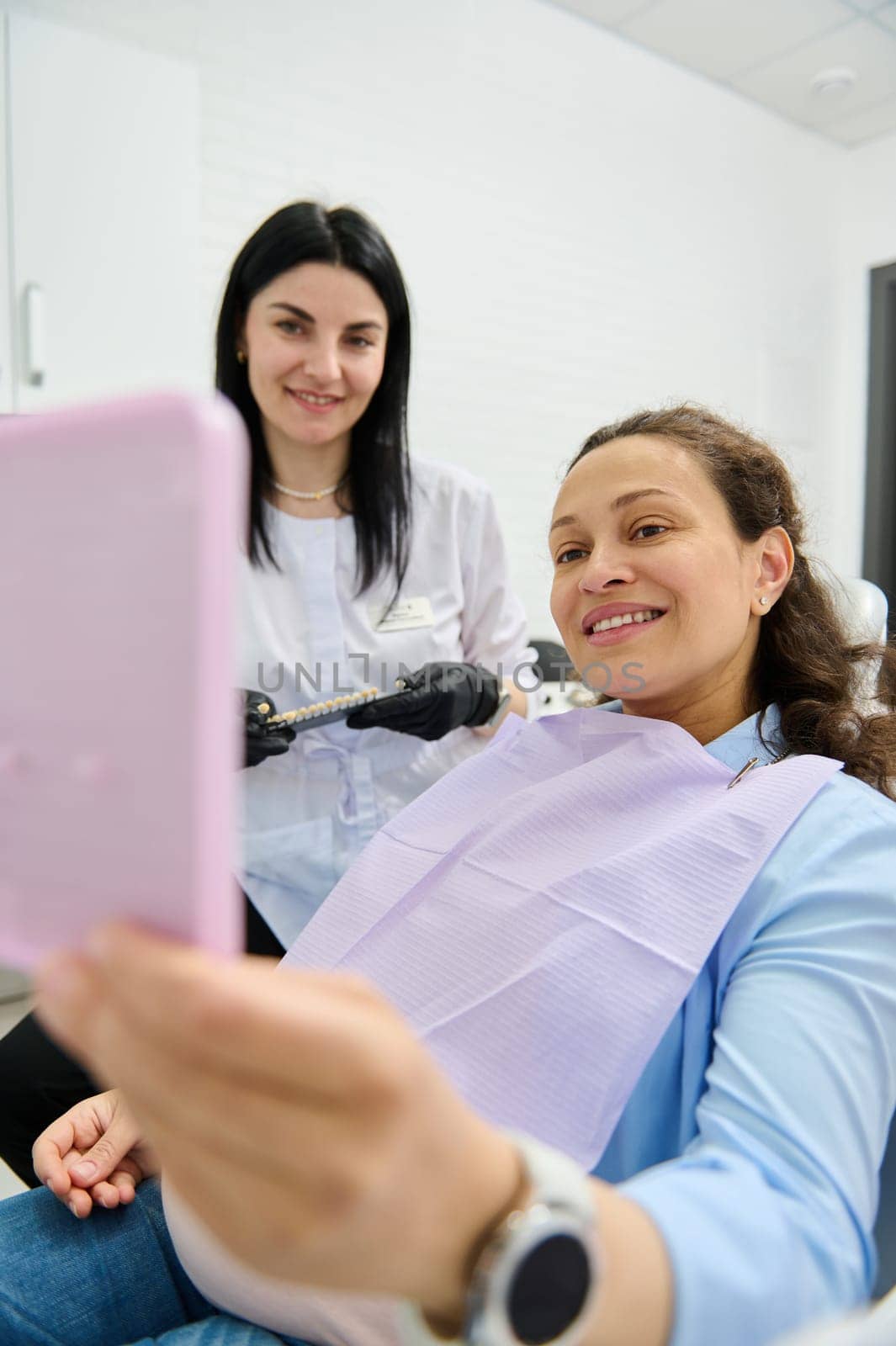 Portrait of a pregnant woman smiling looking at her mirror reflection, sitting in dentist's chair while a dental appointment in a modern dentistry clinic. Dentist holding teeth color chart vita scale