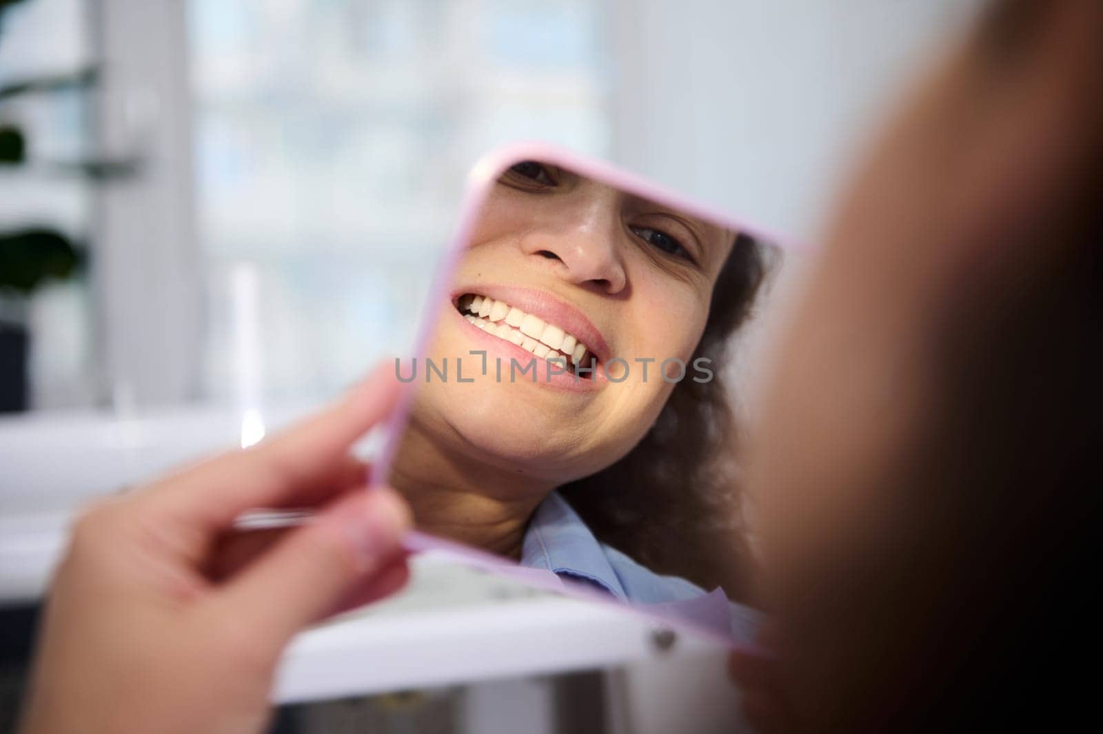 Reflection in mirror of a female patient in dental chair, admiring her smile and teeth after teeth bleaching procedure by artgf