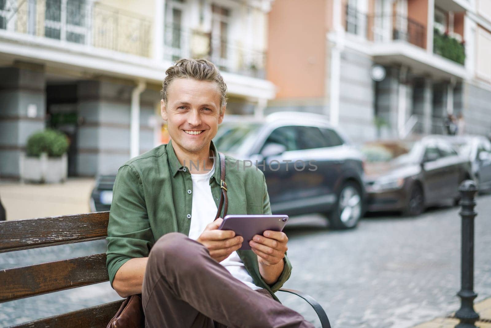 Integration of technology into everyday life, as man utilizes digital devices for information and communication. Blond man searching for information on internet using tablet PC on bench in city street. . High quality photo