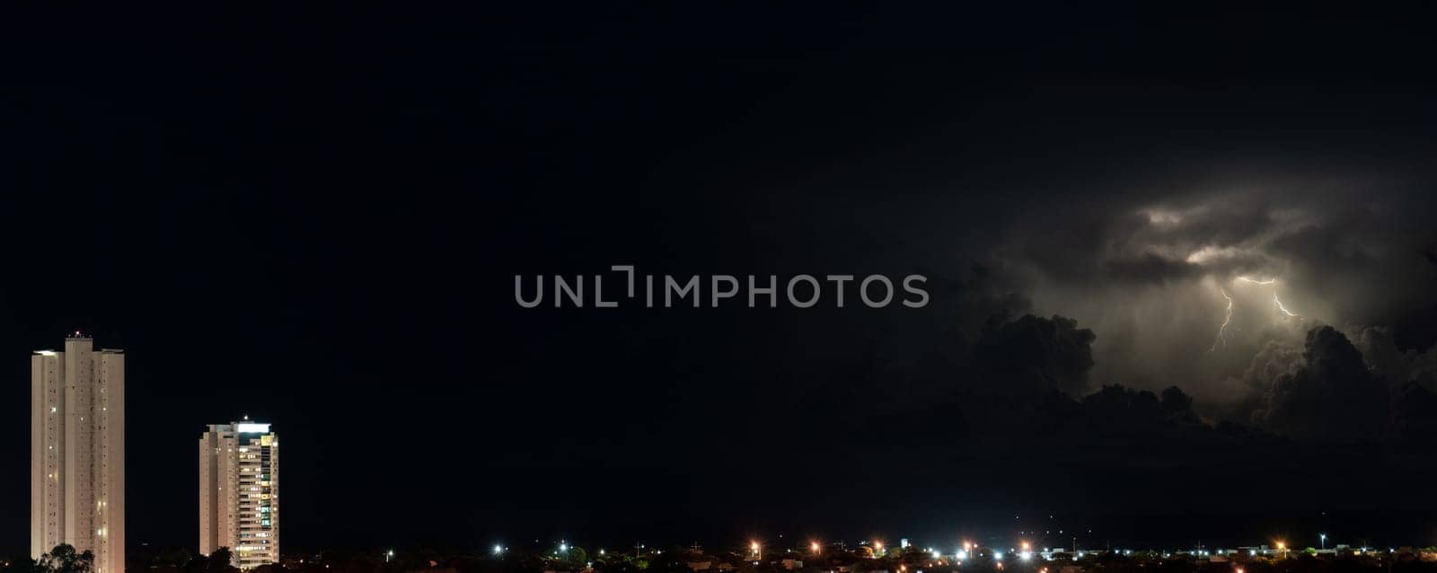 Two skyscrapers rise above the city in a majestic manner during a dark night with thunder and lightning in the background. Space for text available.