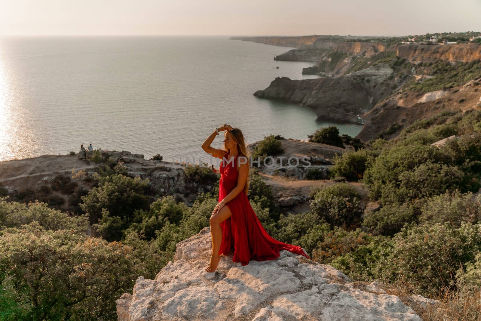 Woman sunset sea red dress, side view a happy beautiful sensual woman in a red long dress posing on a rock high above the sea on sunset. by Matiunina