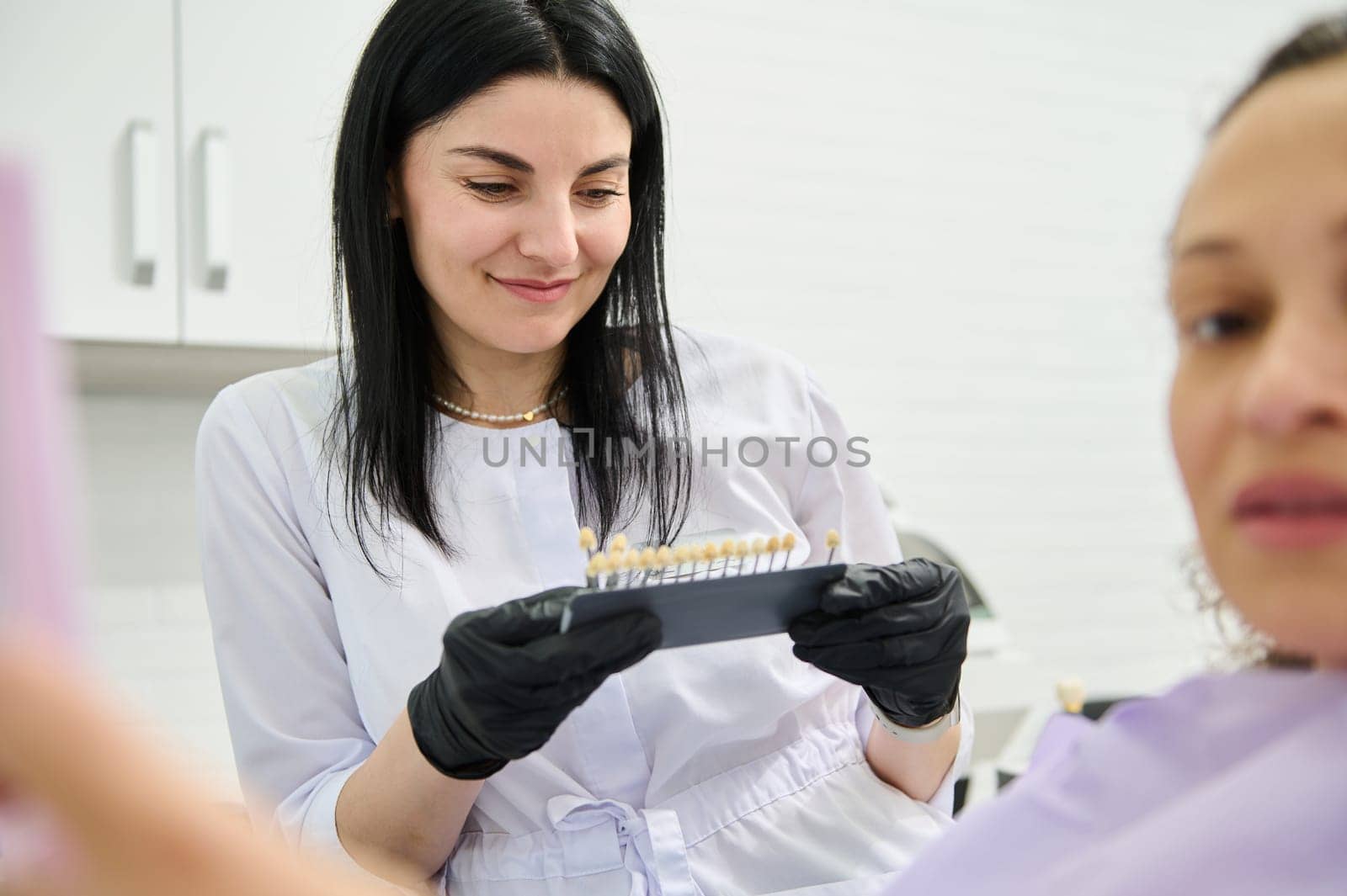 Smiling beautiful woman dentist orthodontist holding teeth color and shade chart guide according to Vita scale. Confident female doctor helping patient to choose shade for teeth bleaching procedure