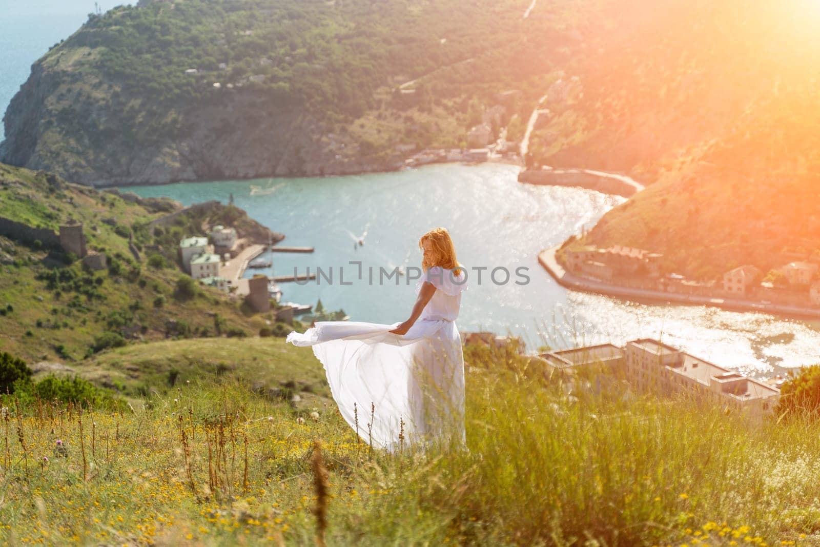 Happy woman in a white dress and hat stands on a rocky cliff above the sea, with the beautiful silhouette of hills in thick fog in the background. by Matiunina