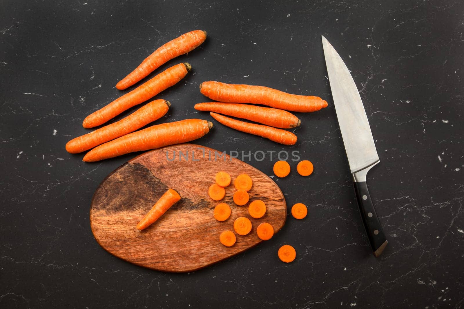 Top down view, chopping board, chef knife and carrot, some of them sliced, on dark marble board.