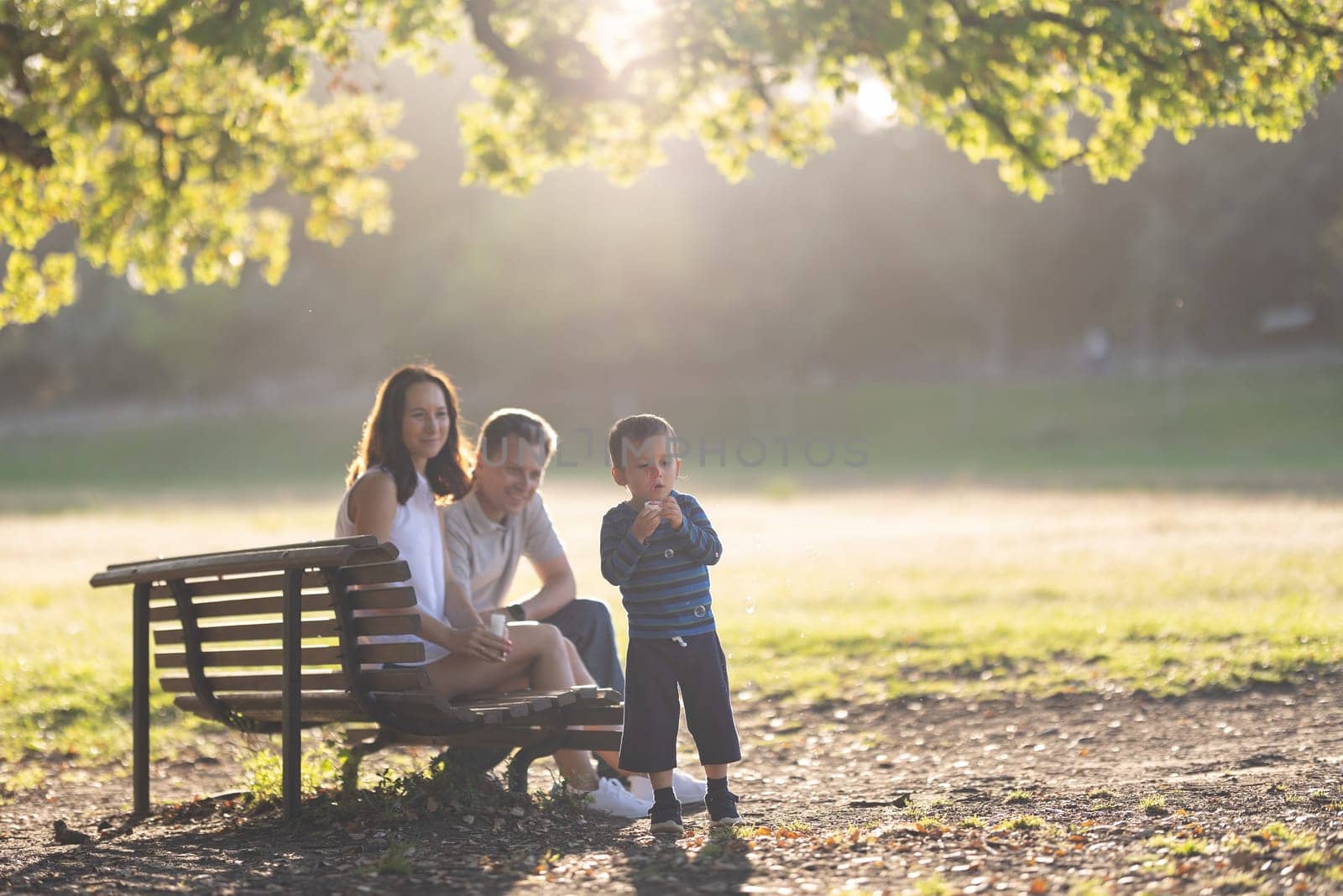 Cute white family in the park - a little boy blowing soap bubbles and his parents watching him. Mid shot