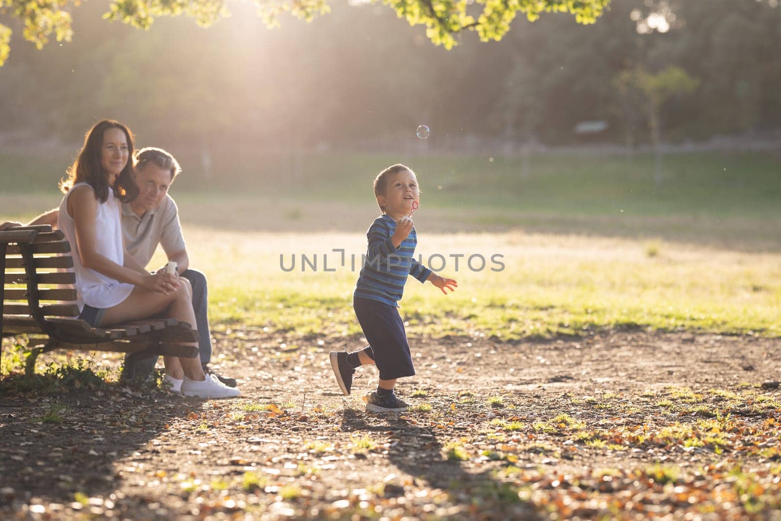 Cute white family in the park - a little boy chasing soap bubbles and his parents watching him. Mid shot