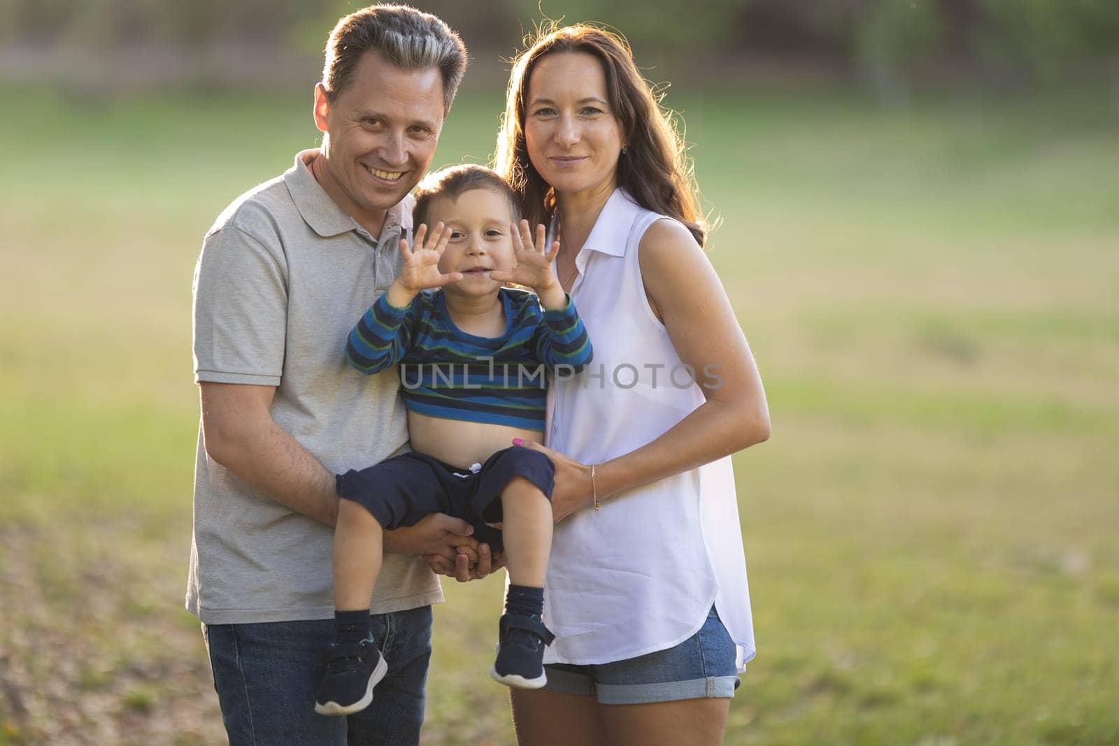 Cute white parents holding their little son on their arms in the sunny park. Mid shot