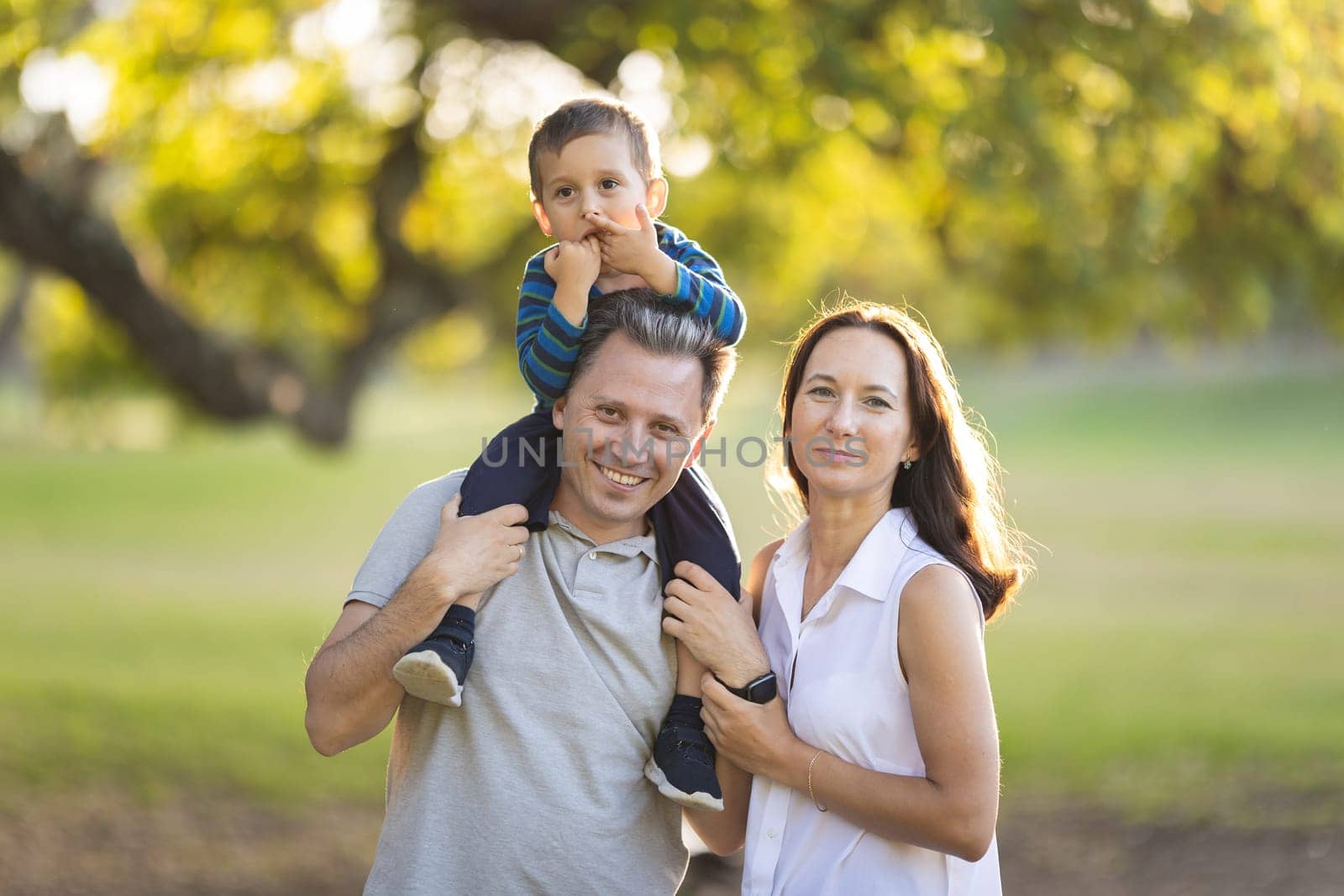 Cute white smiling family in the park - little son on the shoulders of his father and his wife standing by by Studia72