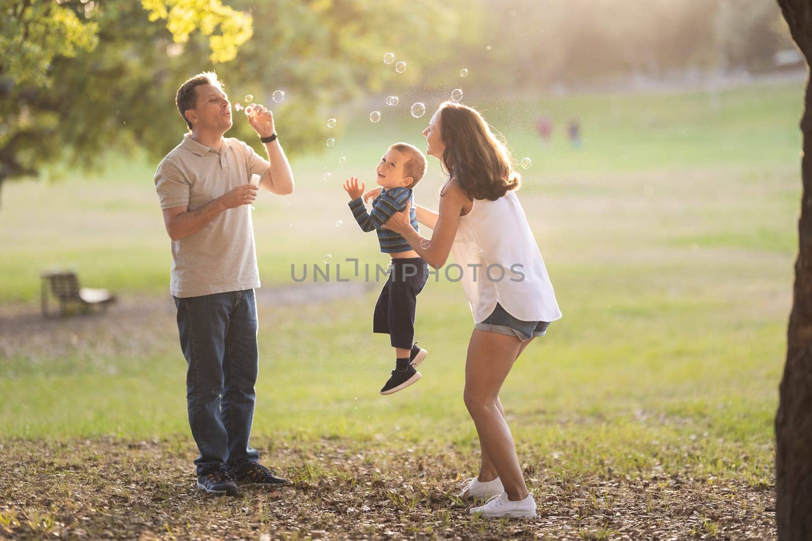 Happy white family spending time in the park - father blowing soap bubbles and his wife and son smiling. Mid shot