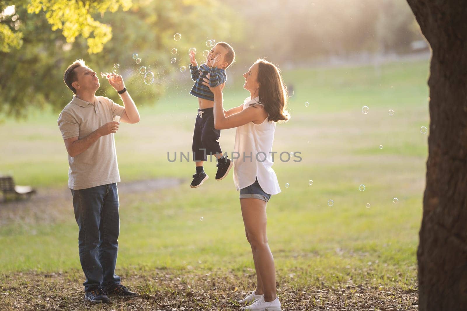 Happy white family spending time in the park - little boy held by his mother catching the soap bubbles his father blowing. Mid shot