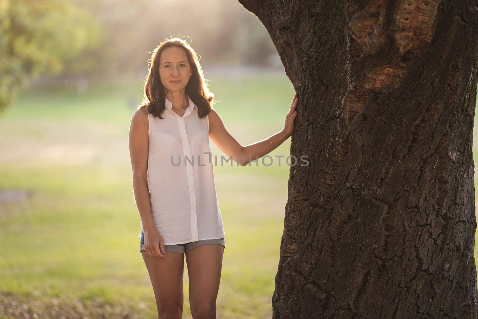 Adult white woman standing by tree trunk. Mid shot