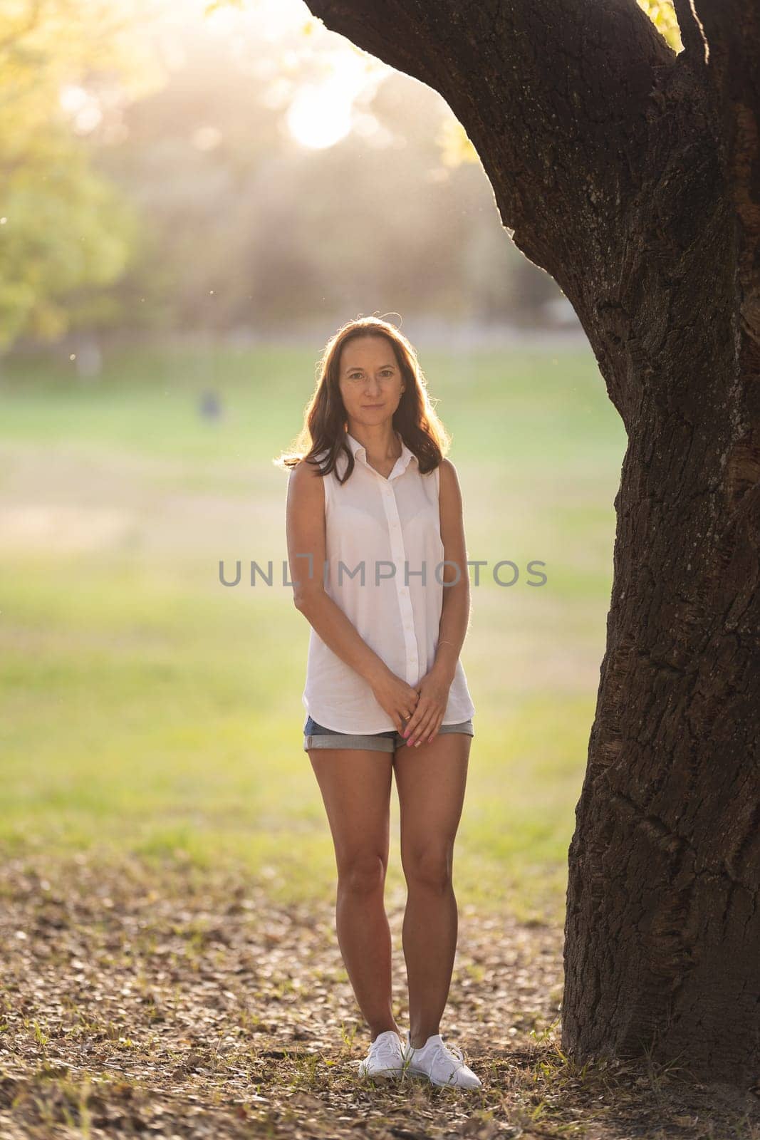 Adult white woman with brown hair standing by tree trunk. Vertical shot