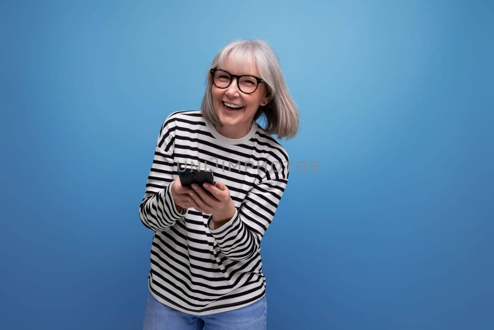 well-groomed slender 60s middle-aged woman holding a smartphone with a mockup on a bright background with copy space.