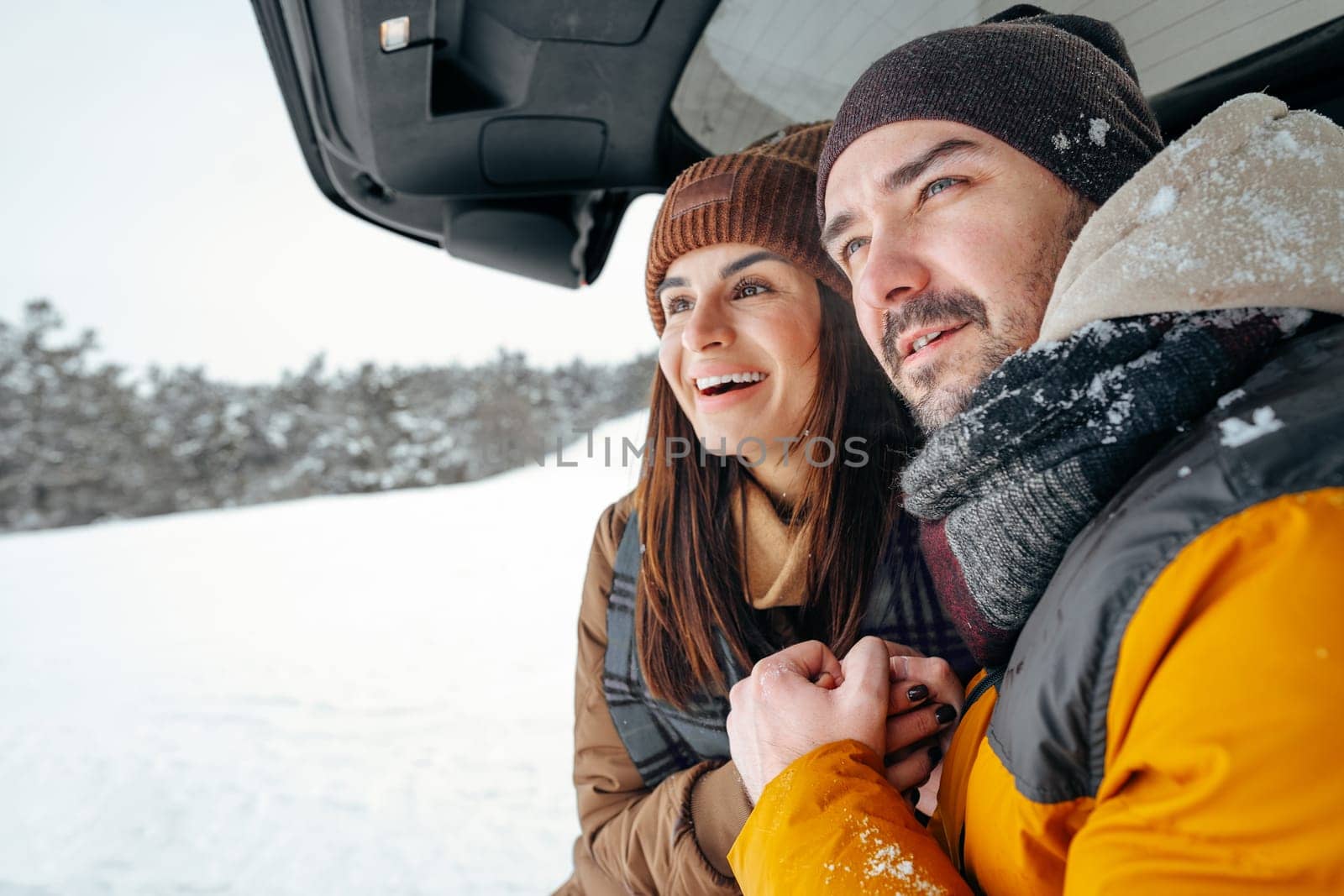 Lovely smiling couple sitting in car trunk in winter forest, close up