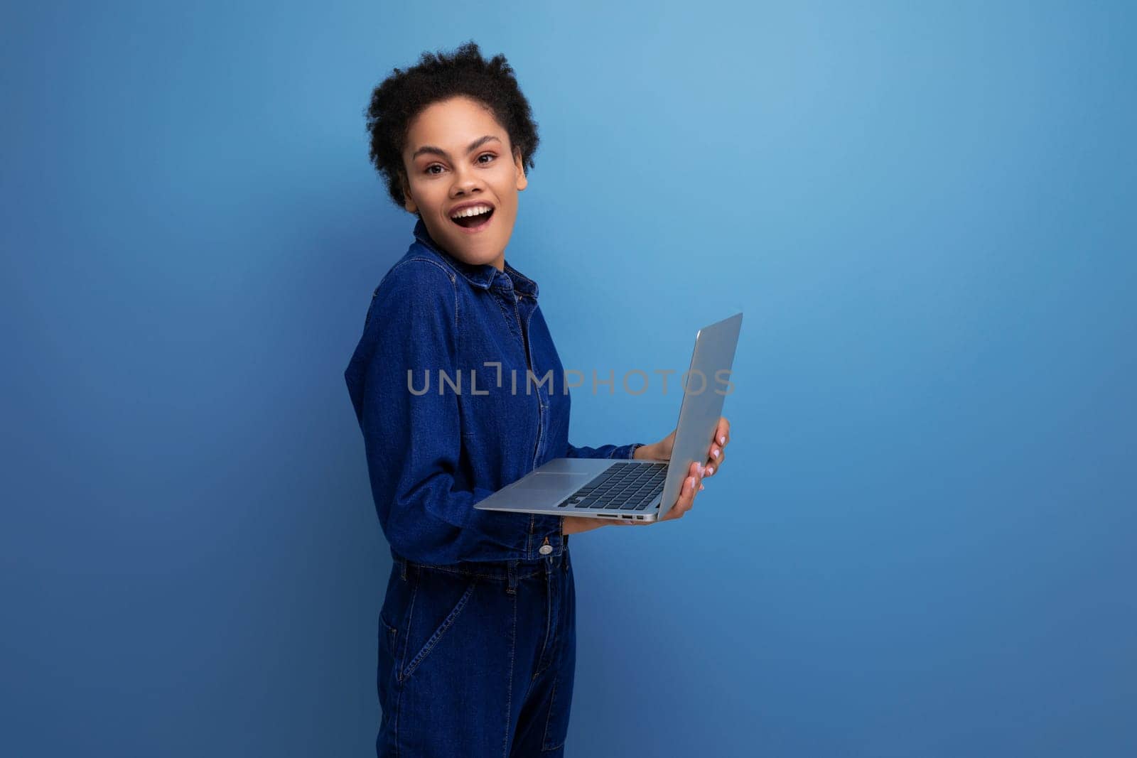 young happy brunette latin woman dressed in blue denim overalls studying remotely as a programmer and holding a laptop in her hands.