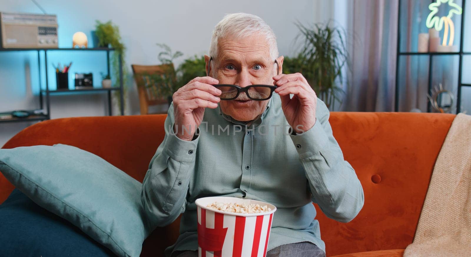 Senior grandfather man in 3D glasses on sofa eating popcorn snacks and watching interesting TV serial sport game, film, online social media movie content at home apartment. Guy enjoying entertainment