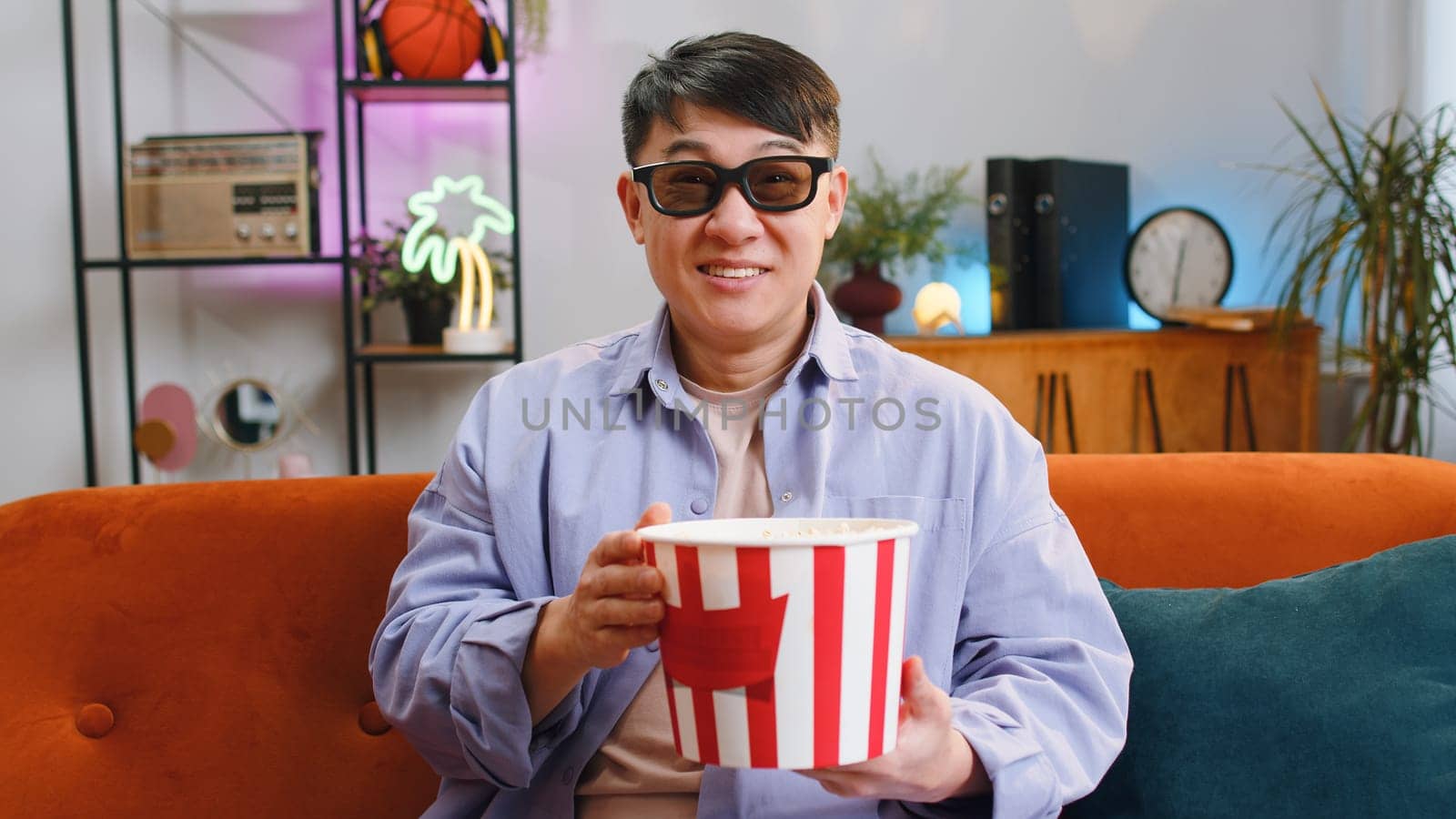 Excited asian man in 3D glasses sits on sofa eating popcorn snacks and watching interesting TV serial, sport game, film, online social media movie content at home apartment. Guy enjoying entertainment
