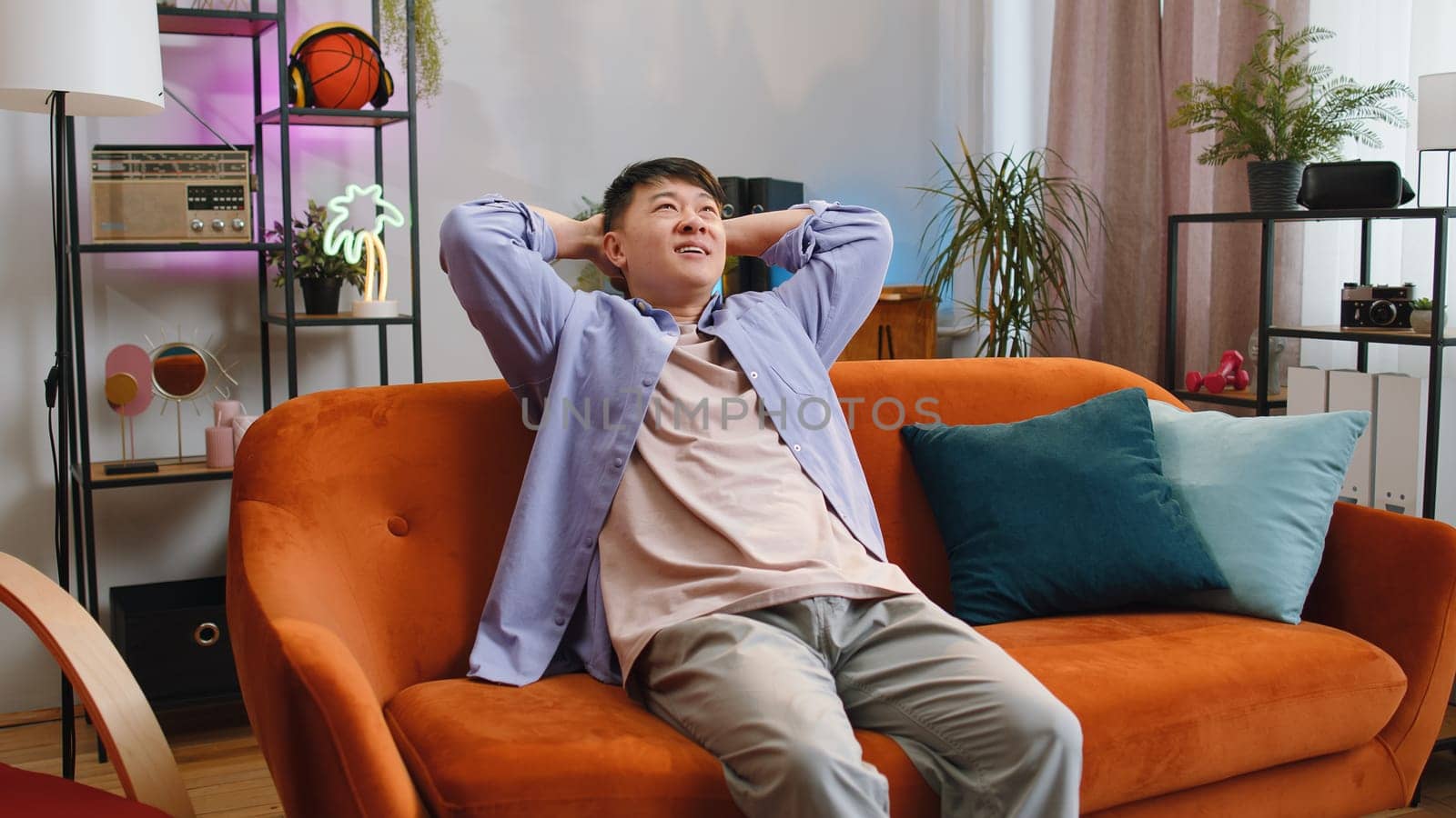 Tired asian handsome man enjoy relaxing on home sofa in living room. Happy chinese guy sits on couch resting napping after hard working day, closed his eyes, put his hands behind his head, stress free