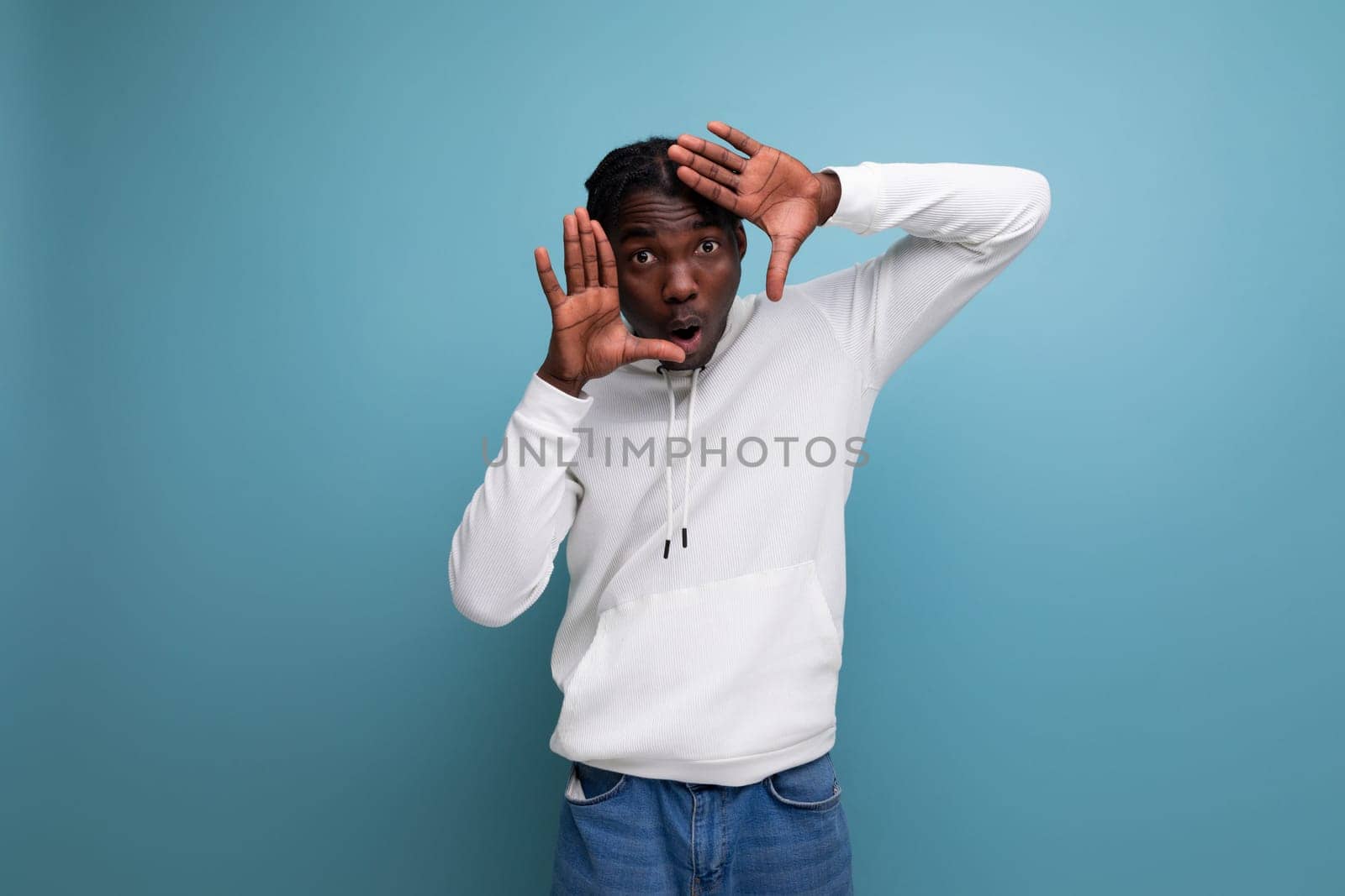 active american young man with dreadlocks in a white sweater inspired shows his hand to the side on a blue background.