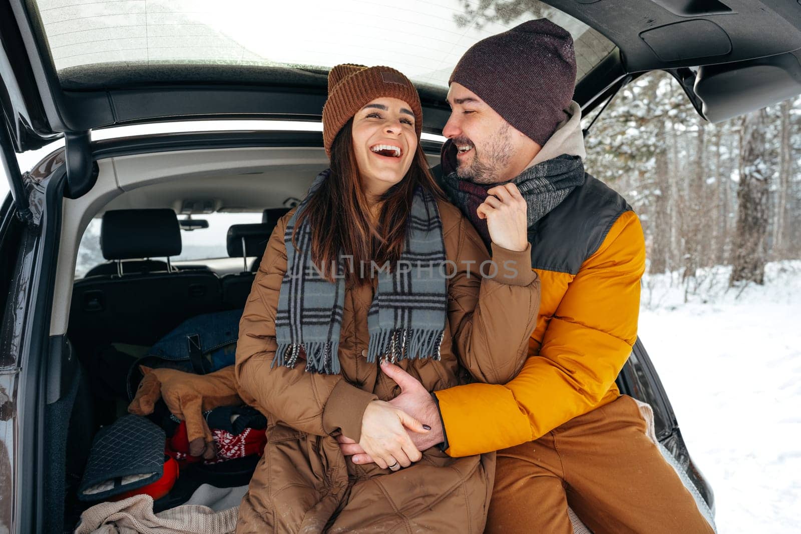 Lovely smiling couple sitting in car trunk in winter forest by Fabrikasimf