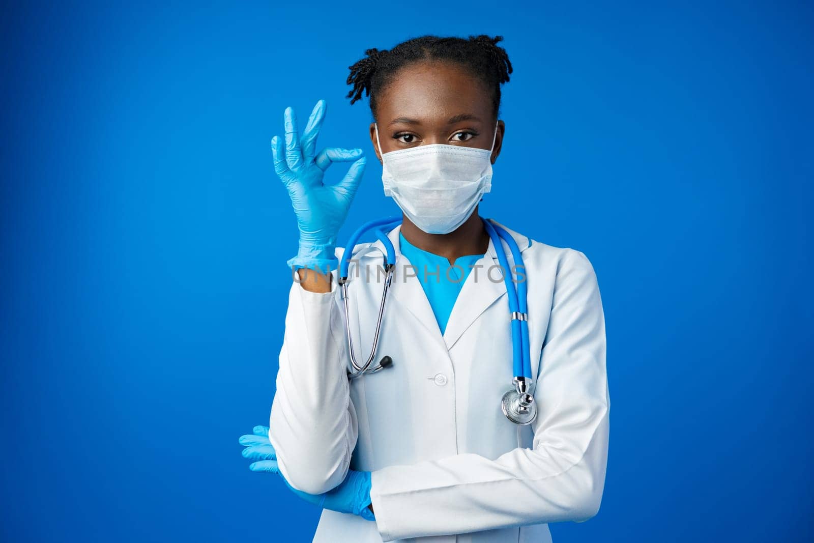 Portrait happy African American female doctor showing ok sign in blue studio by Fabrikasimf