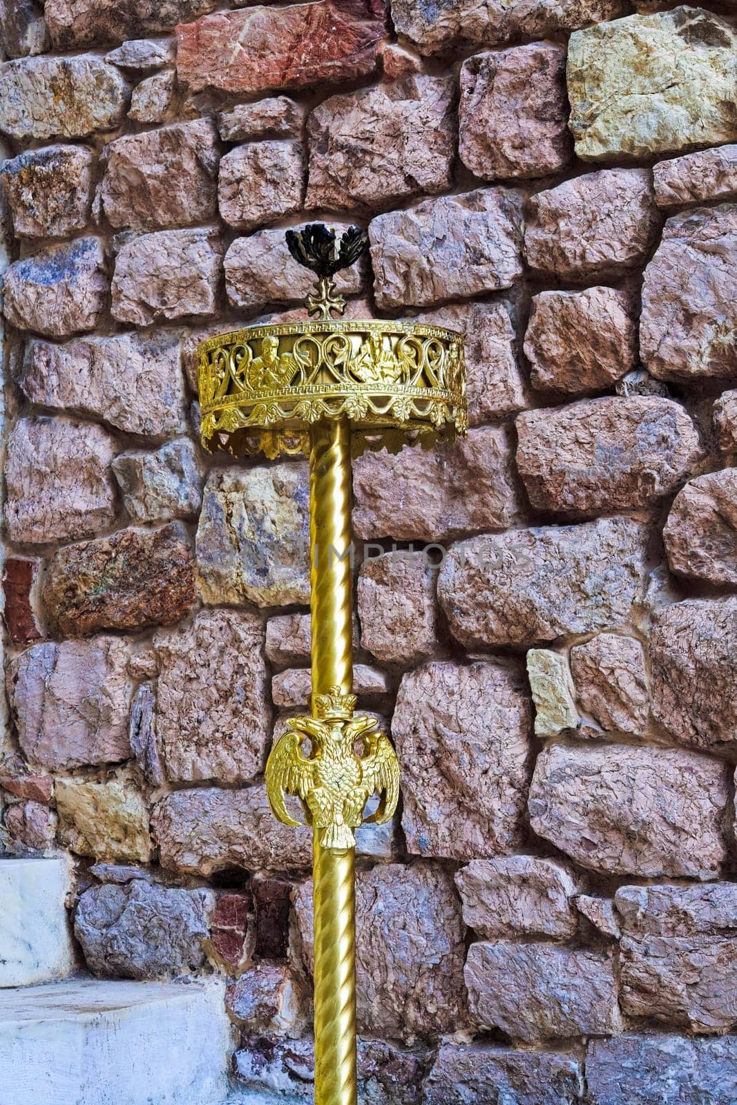A golden candle holder with the double-headed eagle symbol of the Byzantine Empire outside a stone brick wall.