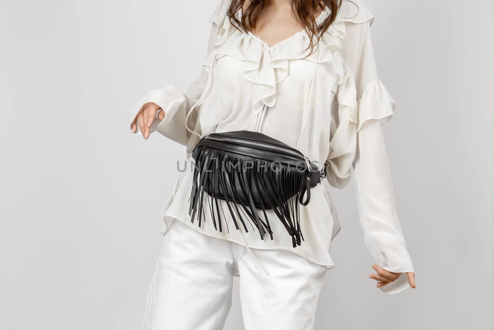 White Woman Wearing White Jeans, Blouse with Frills and Crossbody Black Leather Belt Bag with Decorative Fringe over Grey Background