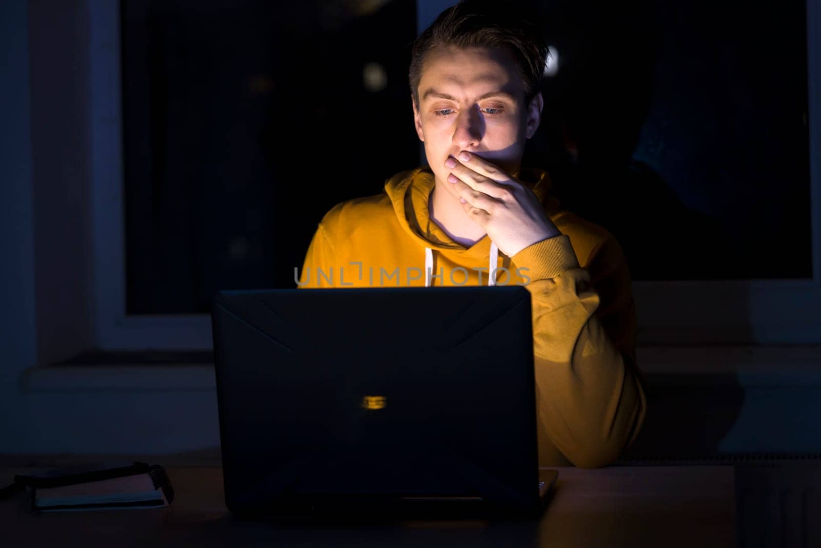A young man sits and works at night on a laptop on the network, types on the keyboard, writes text messages, studies, prepares a project, a programmer, web designer works as a freelancer at home.