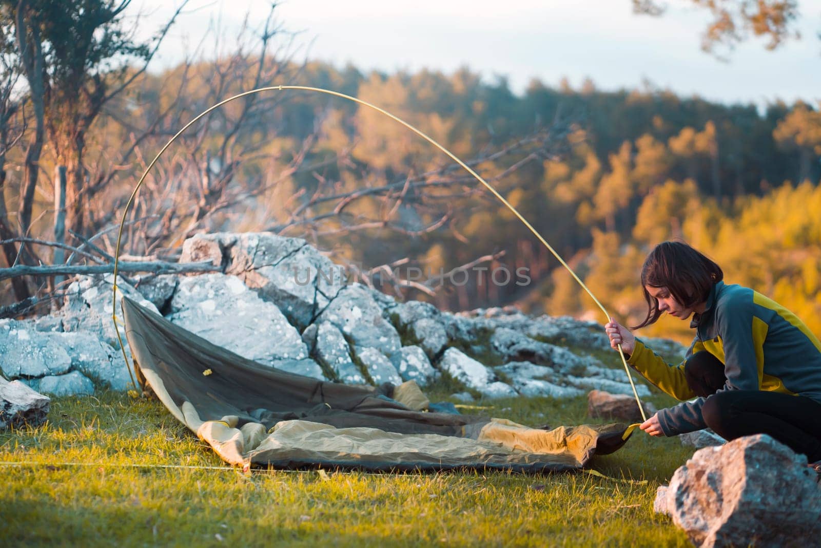 A young girl sets up her tent at a campsite on a green meadow among the mountains on a warm summer evening. The traveler is getting ready for a comfortable sleep in nature.