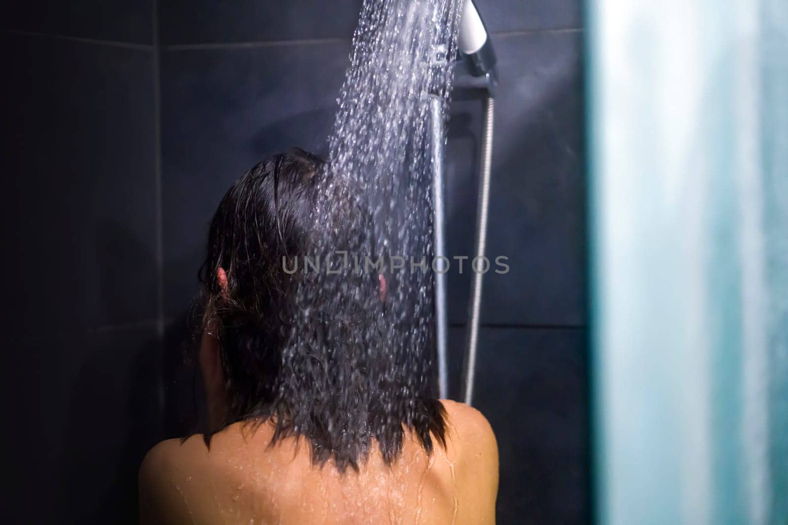 A young girl takes a shower with shampoo and soap, washes her hair, takes care of her body, hygiene and cleanliness in a modern gray shower room.
