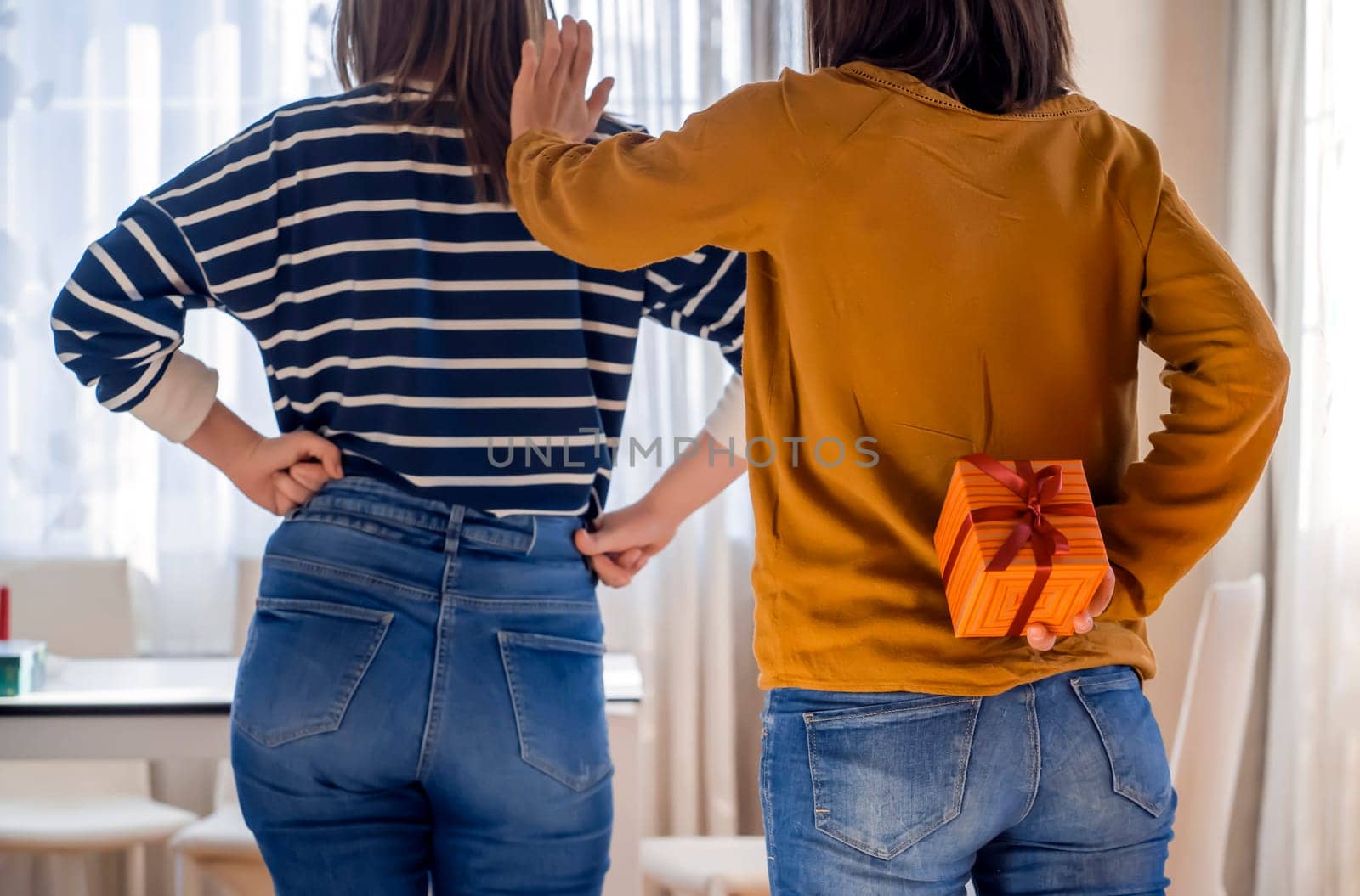 Funny girl tiptoes up to her friend with a desire to make a pleasant surprise, hides a cute nice wrapped gift behind her back. Two girls have fun and celebrates holidays, presenting gifts each other.