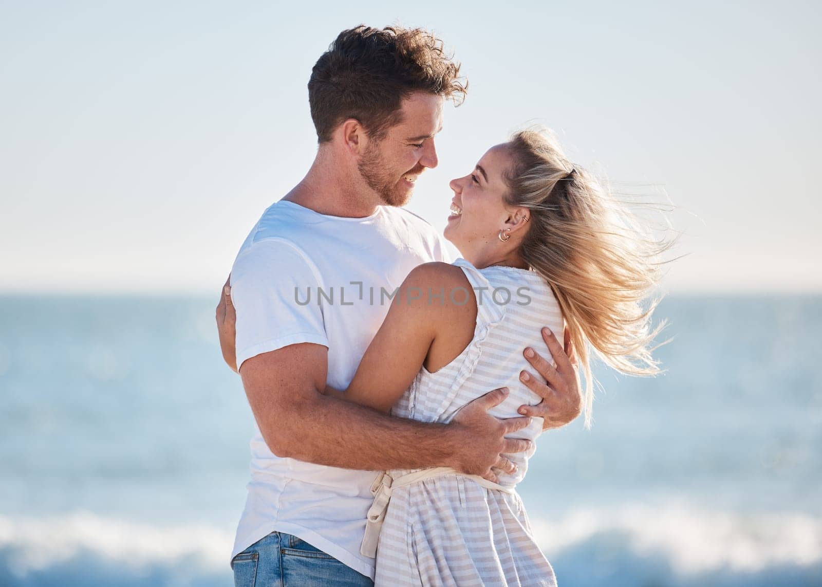 Love, beach hug and couple smile together at the ocean for peace, relax in nature and romance vacation happiness. Happy man, laughing woman and relationship bliss on a travel holiday by sea.