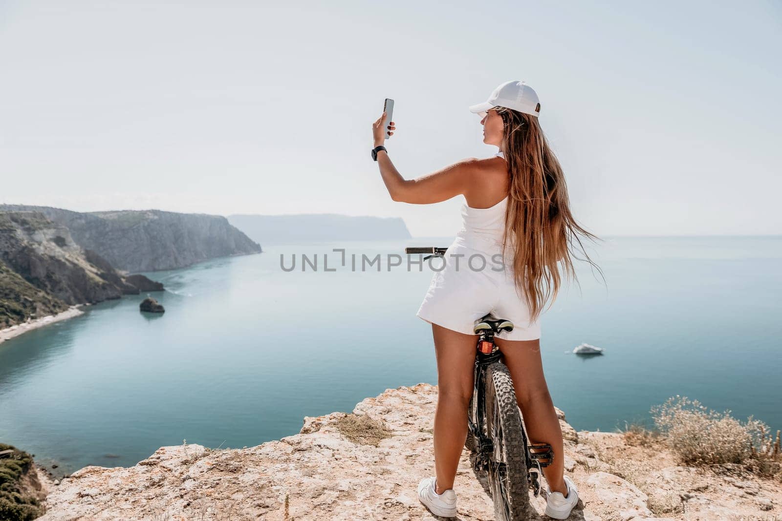 A woman cyclist on a mountain bike looking at the landscape of mountains and sea. Rear view of cyclist woman standing in front to the sea with outstretched arms. Freedom and healthy lifestyle concept