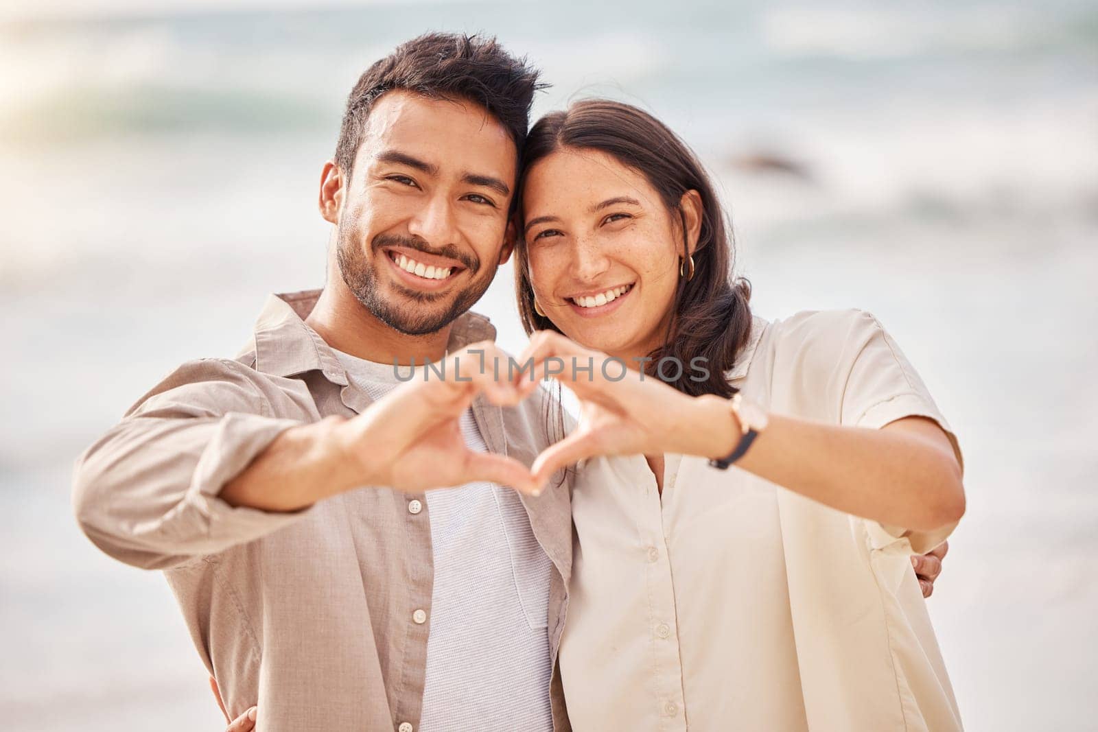 She completes me. Couple, portrait and love sign with hands at the ocean for travel with sunshine with a smile. Beach, together and heart with hand and smile in the summer for a weekend with romance