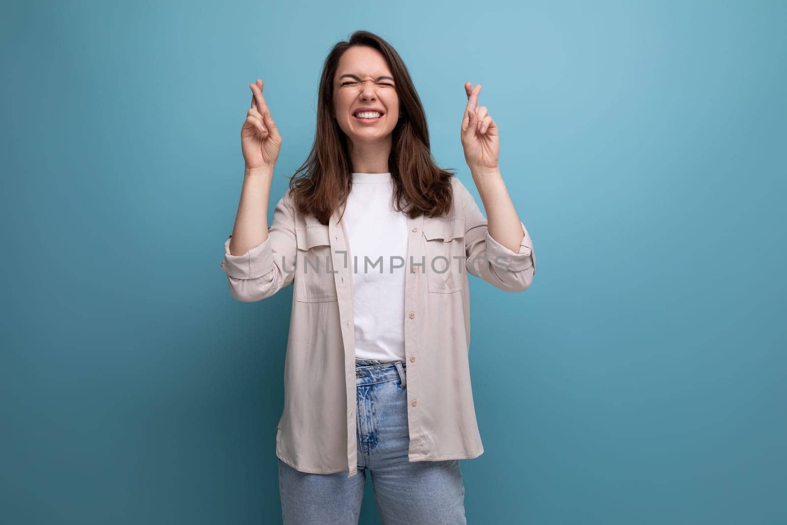 pretty brunette young female adult in shirt and jeans crossed fingers on blue background.