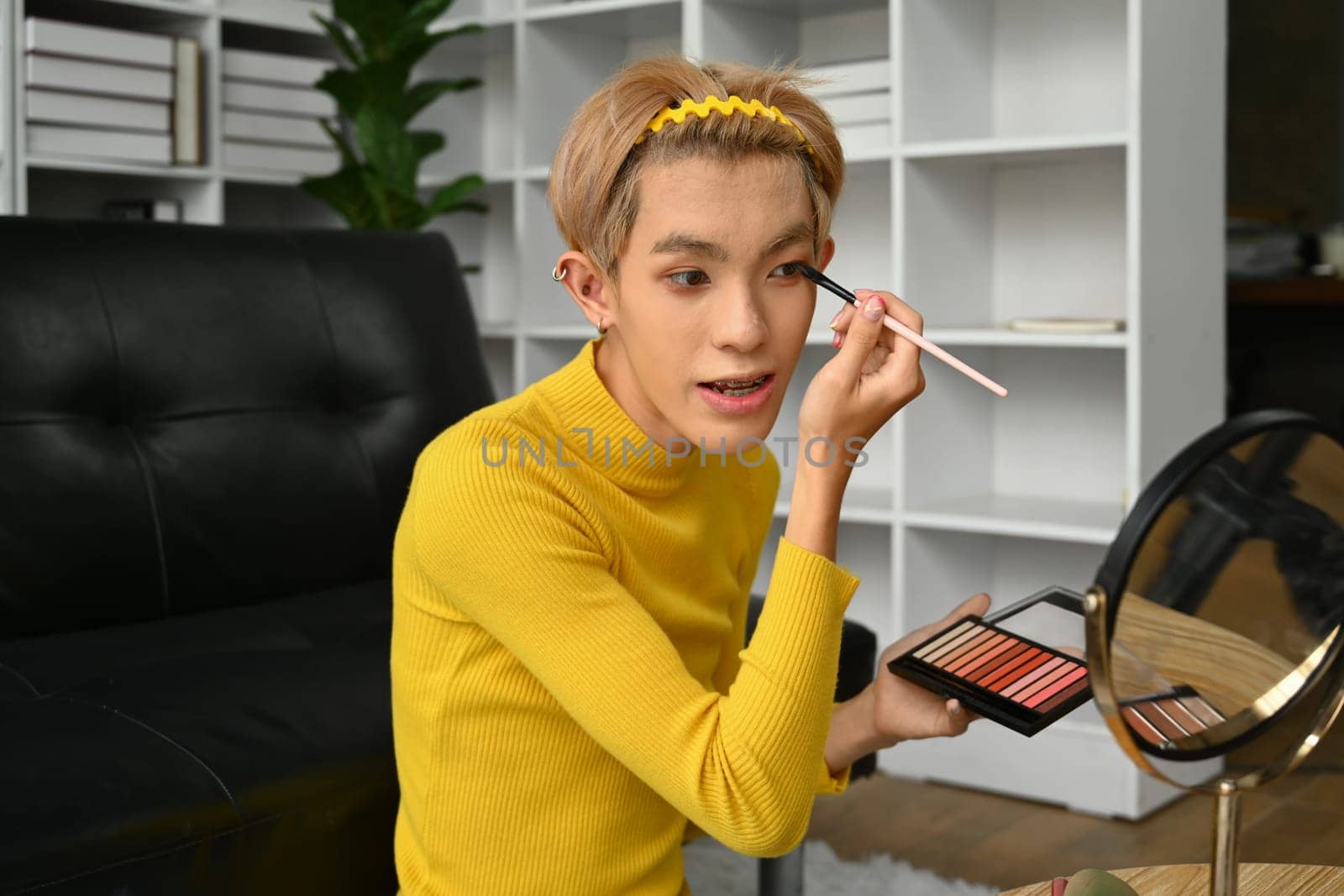 Attractive young gay man looking at mirror and applying eye shadow with brush. LGBTQ, domestic life, fashion and beauty concept.