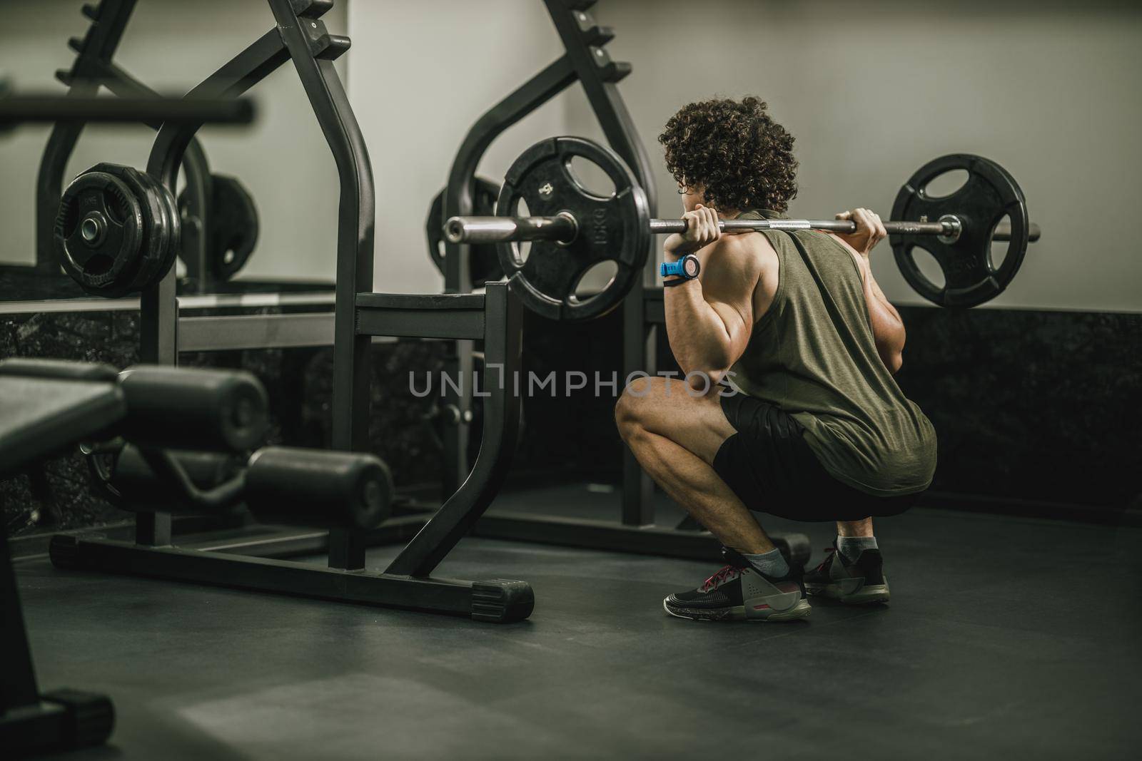 Shot of a muscular guy in sportswear working out at the hard training in a gym. He is doing squat exercises with barbell.