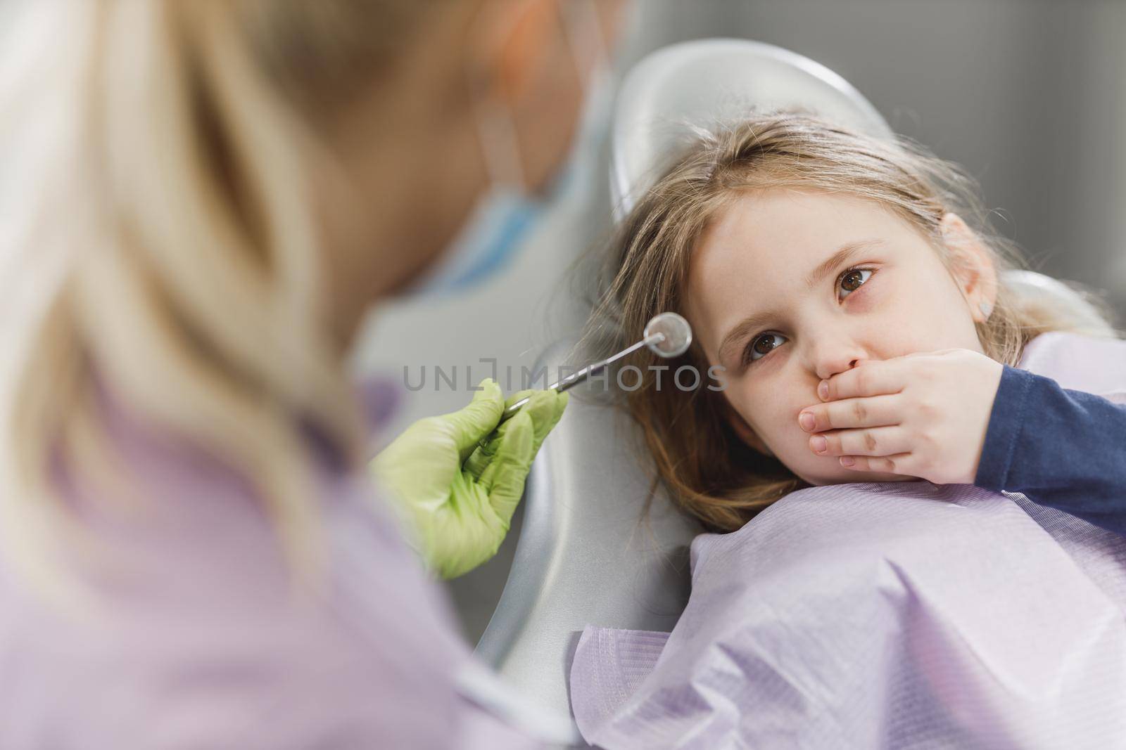 A frightened little girl on a dentist chair and holds her mouth closed to keep the dentist from working.