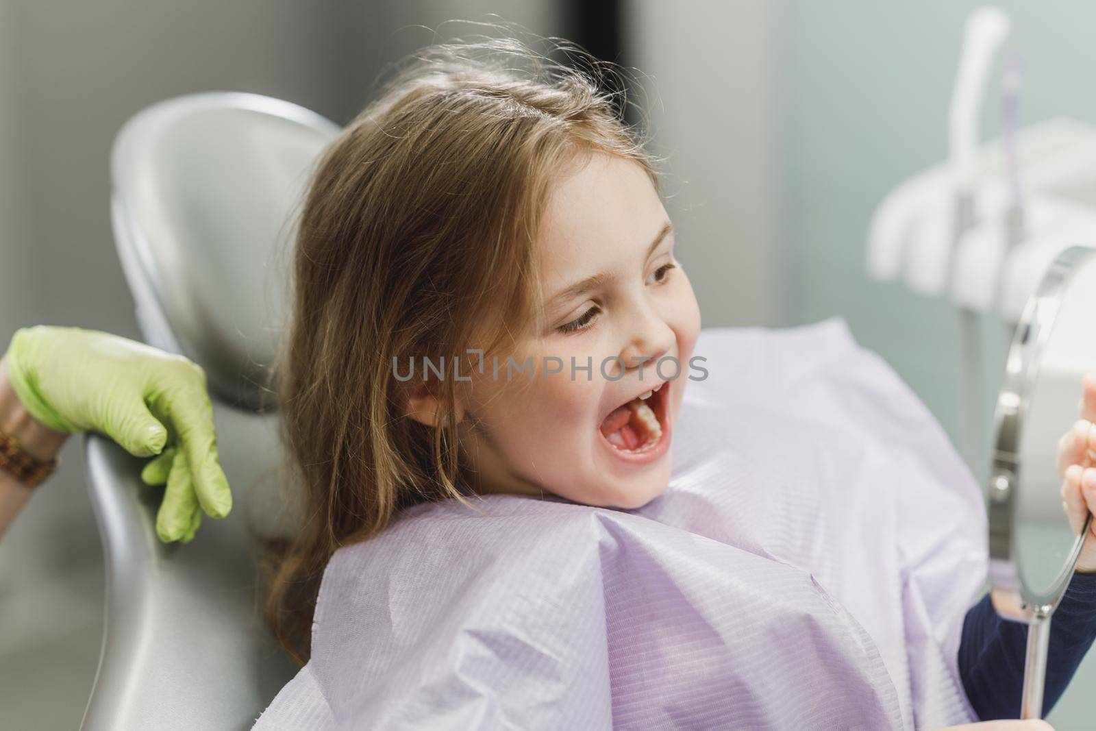A cute little girl looking her teeth in a mirror after dental procedure at dental clinic.