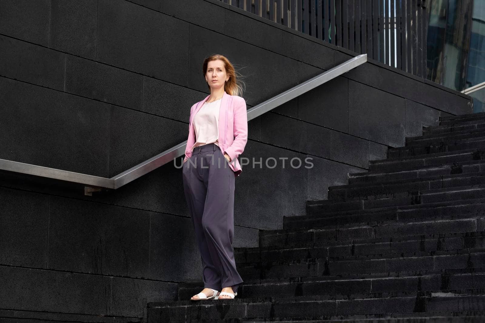 Young serious beautiful blonde millennial woman with long hair in pink elegant clothes stands on steps near modern building. Selective focus.