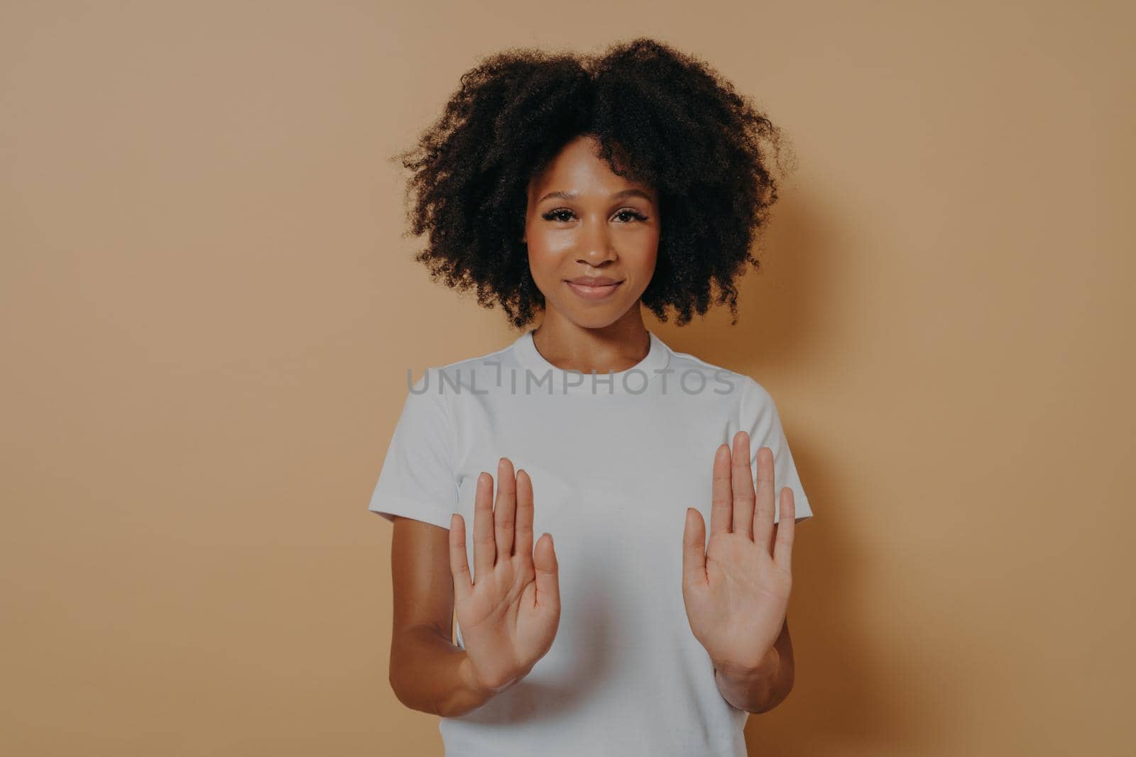 Slow down. Young smiling mixed race woman with curly hairstyle raising palms in stop or prohibition gesture and saying no while standing dressed in white t-shirt over brown studio wall
