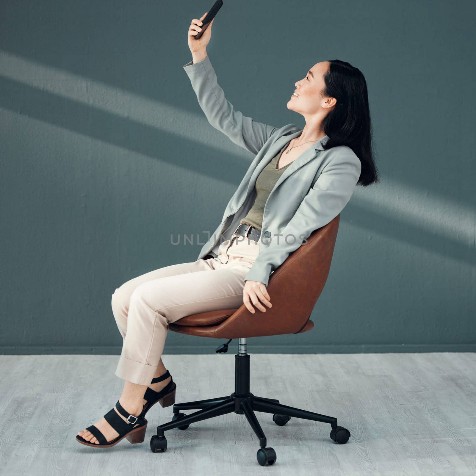 Selfie, video call and woman with a phone for connection, internet and communication at work on a chair. Corporate and Asian employee reading an email, message or chat on a mobile app in an office by YuriArcurs