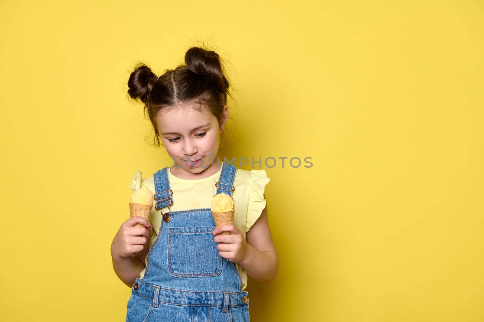 Beautiful baby girl with trendy hairstyle, showing tongue while licking delicious ice cream, isolated on yellow backdrop by artgf