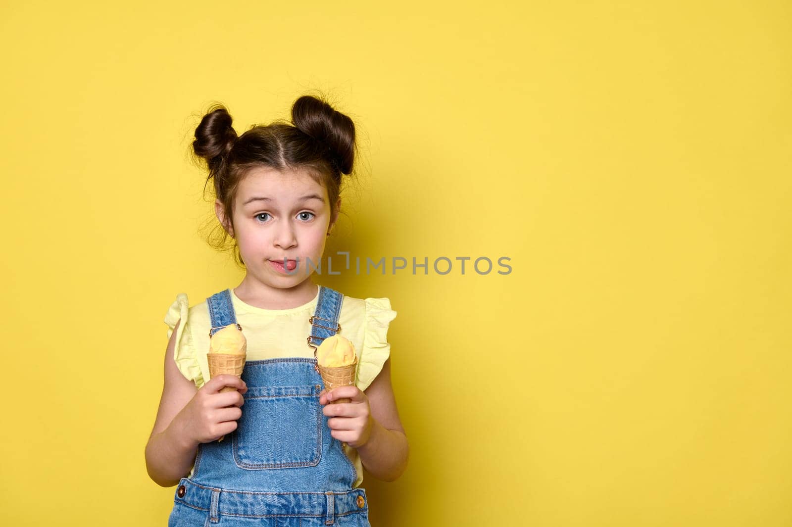 Happy little child girl showing her tongue looking at camera, holding yummy ice cream in waffle cones, popsicle, isolated yellow background with free advertising space for text. Kids. Summer. Children