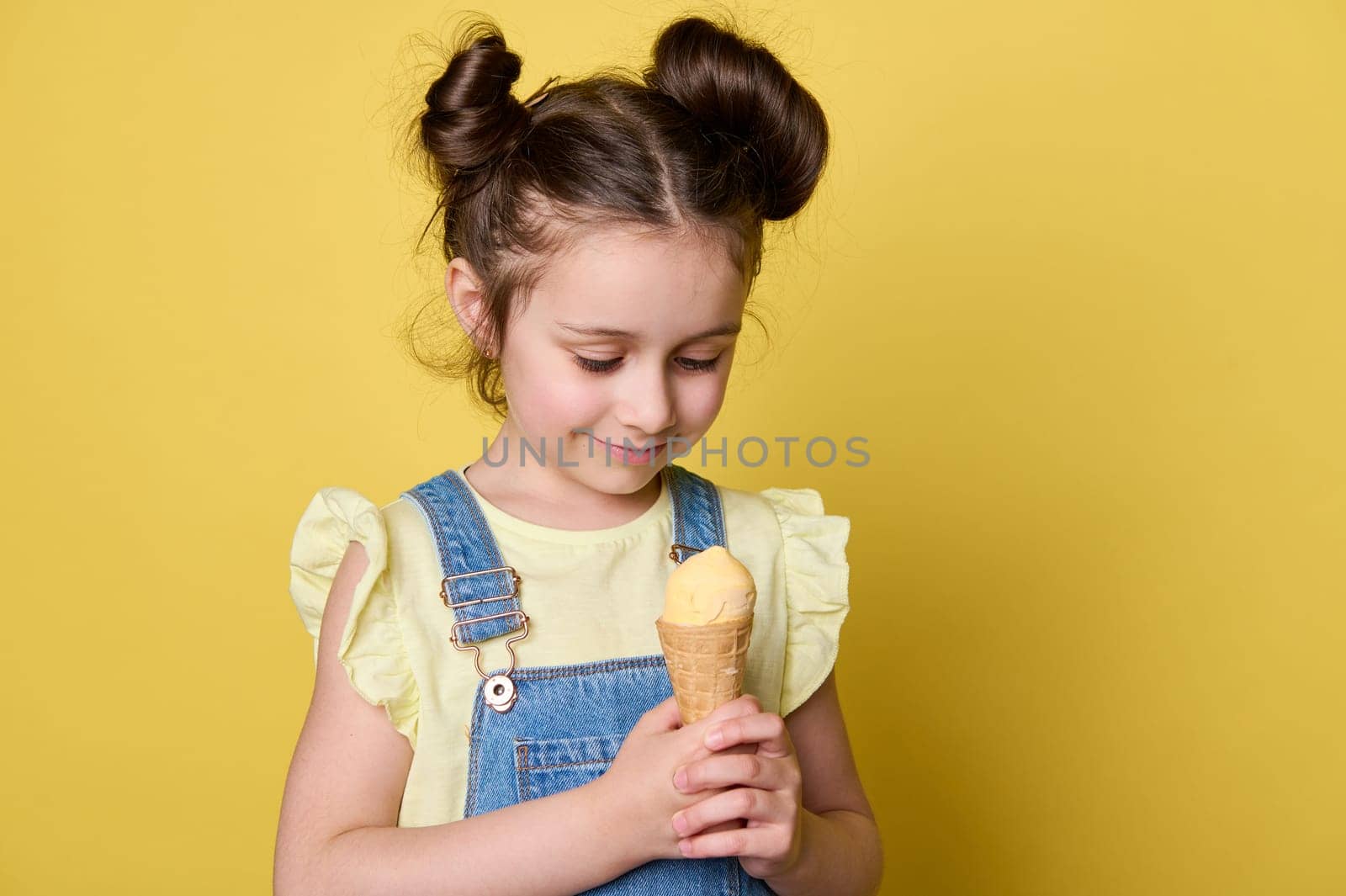 Happy little girl with stylish hairstyle, wearing blue denim overalls and yellow t-shirt, enjoying sweet summer dessert, holding waffle cone with ice cream scoop, smiling isolated on yellow background