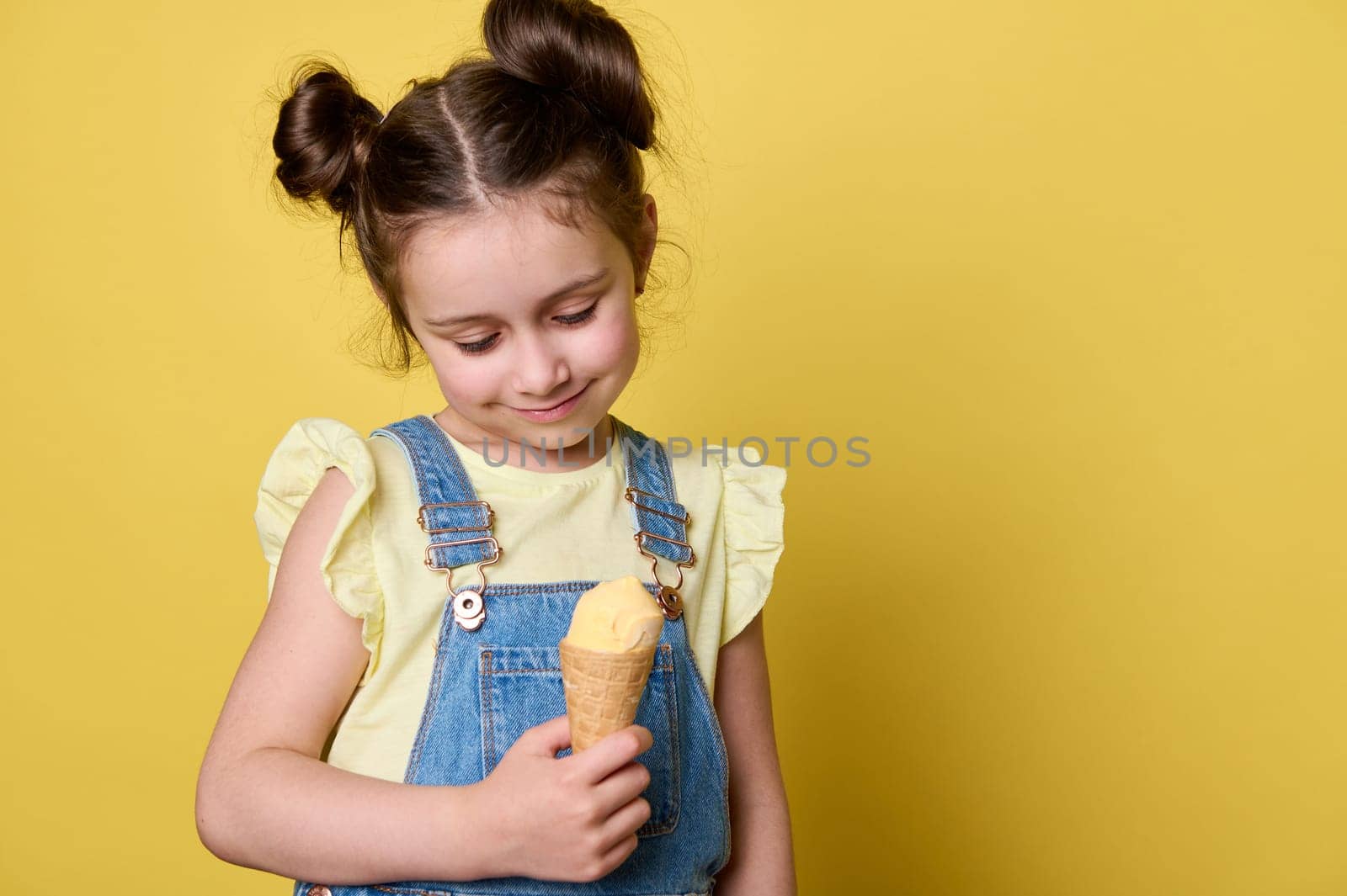 Sweet little girl smiling, holding a waffle cone with a scoop of banana ice cream, isolated on yellow color background by artgf