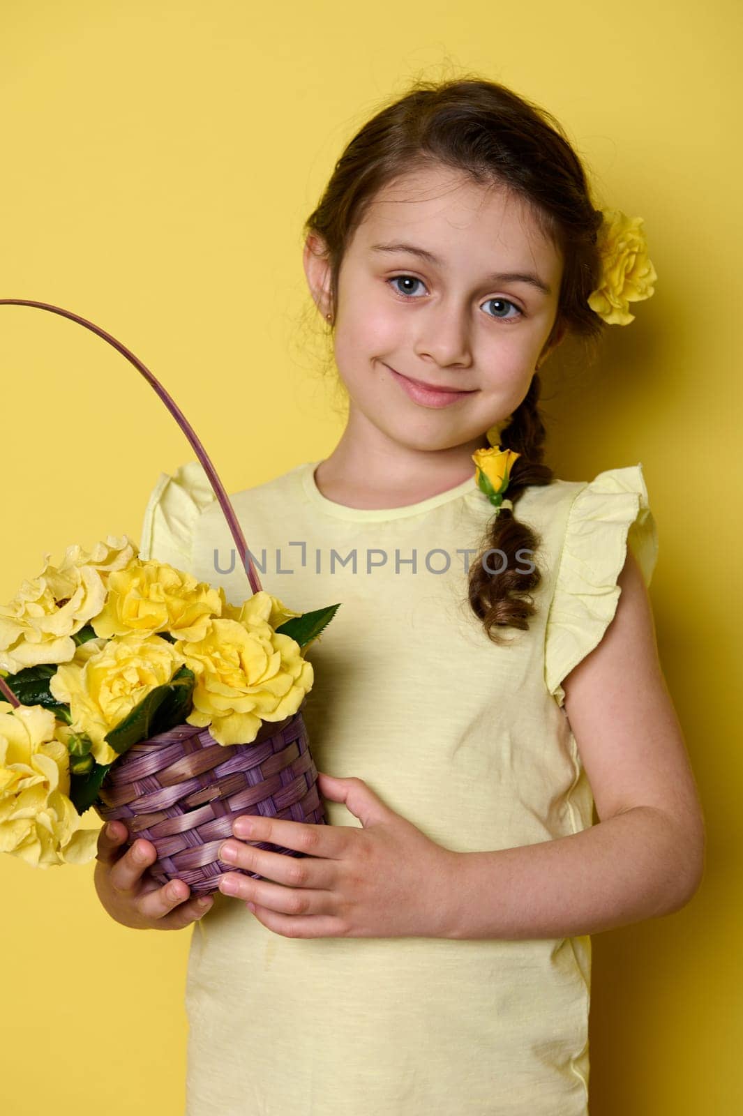 Happy Mother's Day. International children's Day concept. Beautiful lovely child girl, pretty daughter with purple wicker basket of yellow roses, smiling looking at camera, isolated yellow background