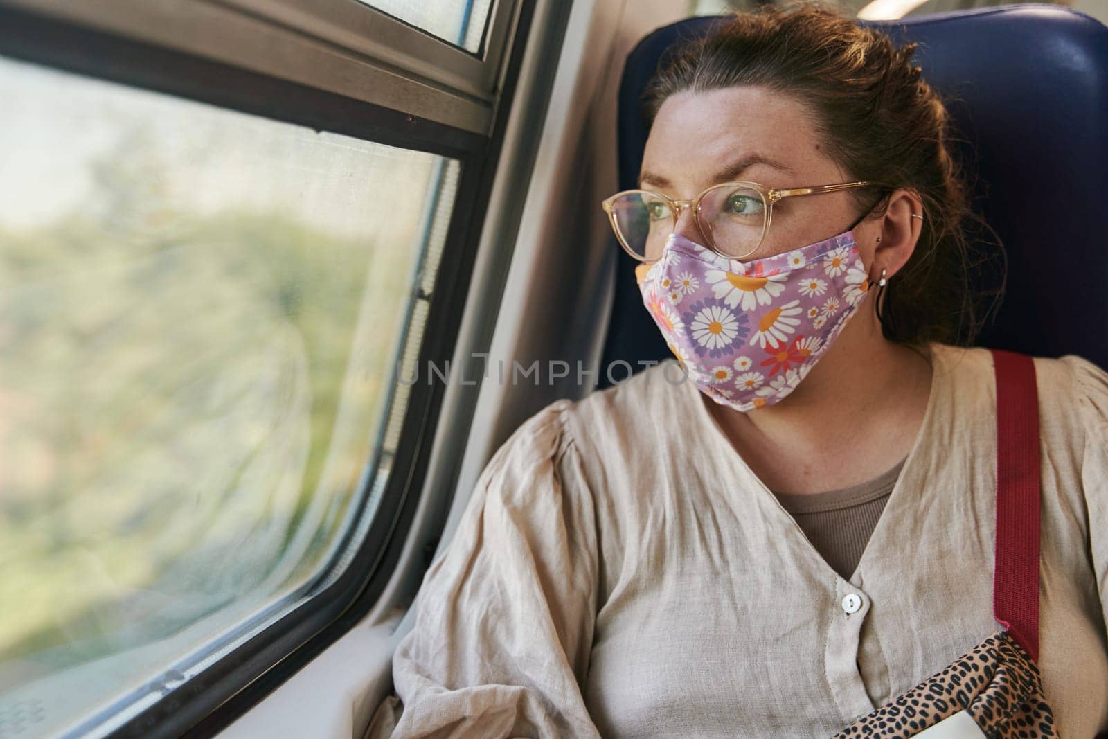 A girl in glasses and a medical mask riding a train and looking out the window. High quality photo